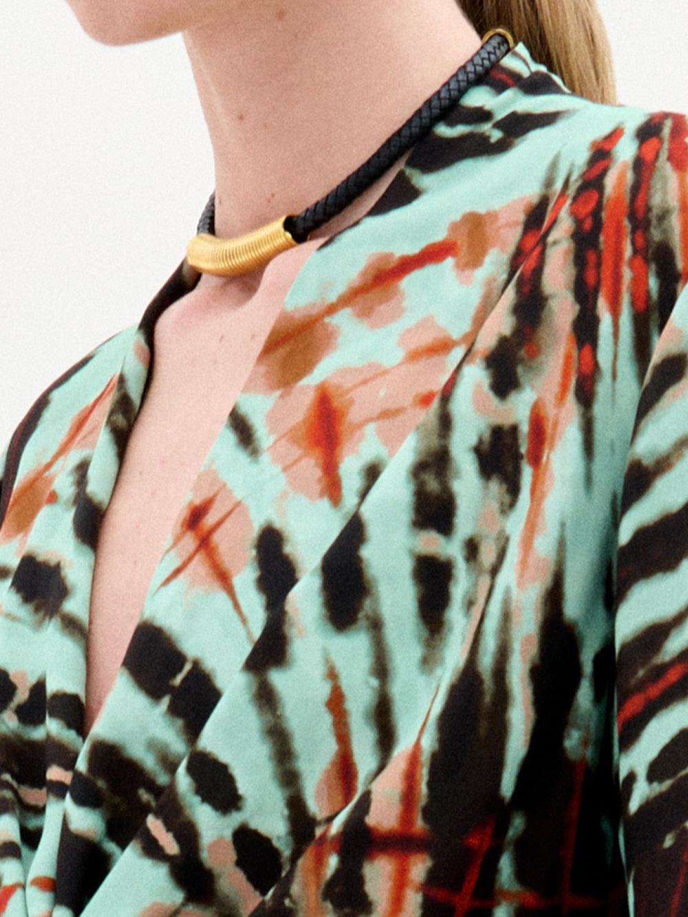 A person wears the Hadria Necklace by SVT DESIGNS—a short leather necklace with a gold curved accent—paired with a colorful turquoise, orange, and black garment. Only their neck, shoulder, and upper chest are visible. Made in Colombia.