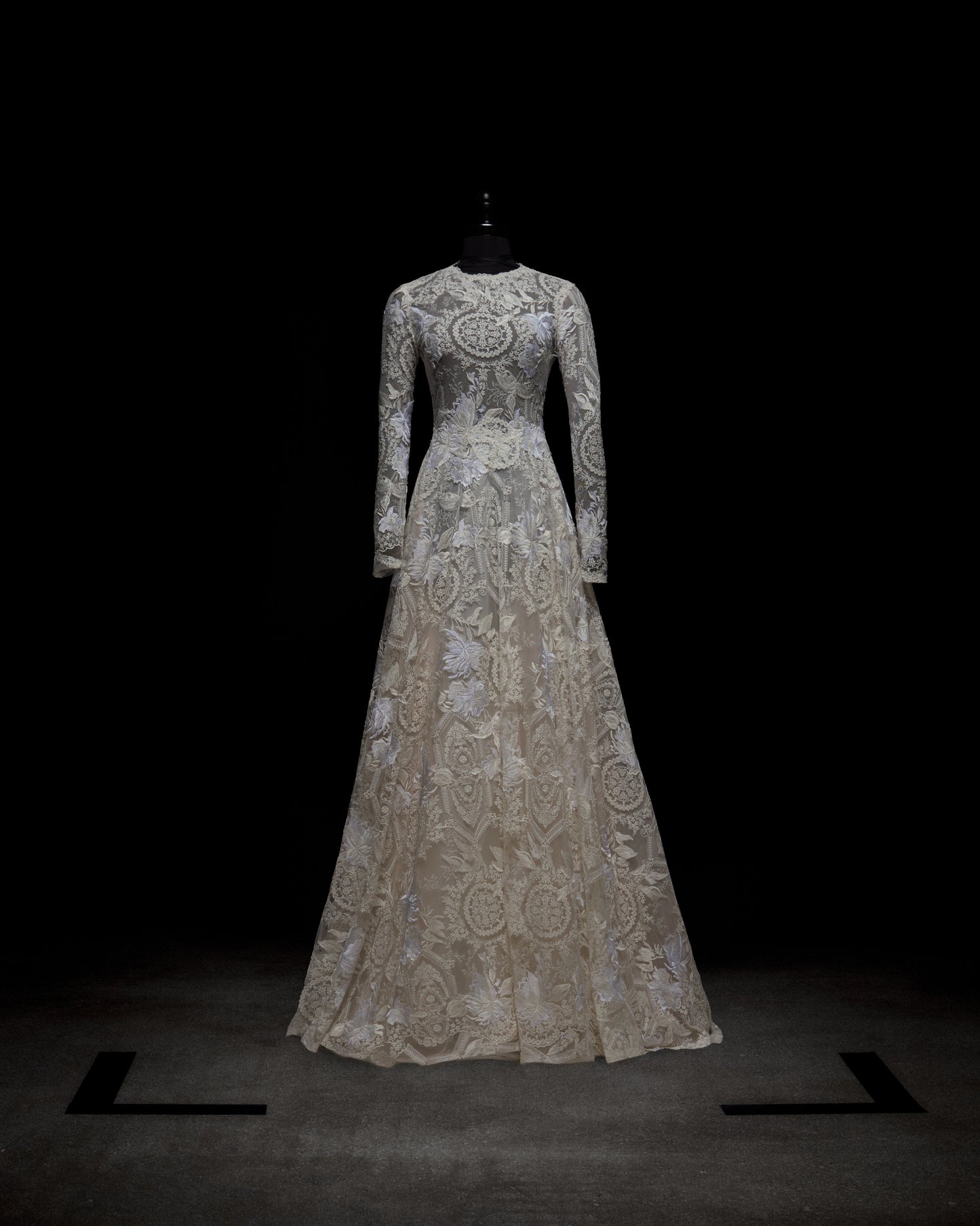 The Hermione Dress by SVT DESIGNS is showcased on a black mannequin against a dark background. It features intricate lace patterns on its long sleeves and fitted bodice that beautifully lead to a flowing, full-length skirt.