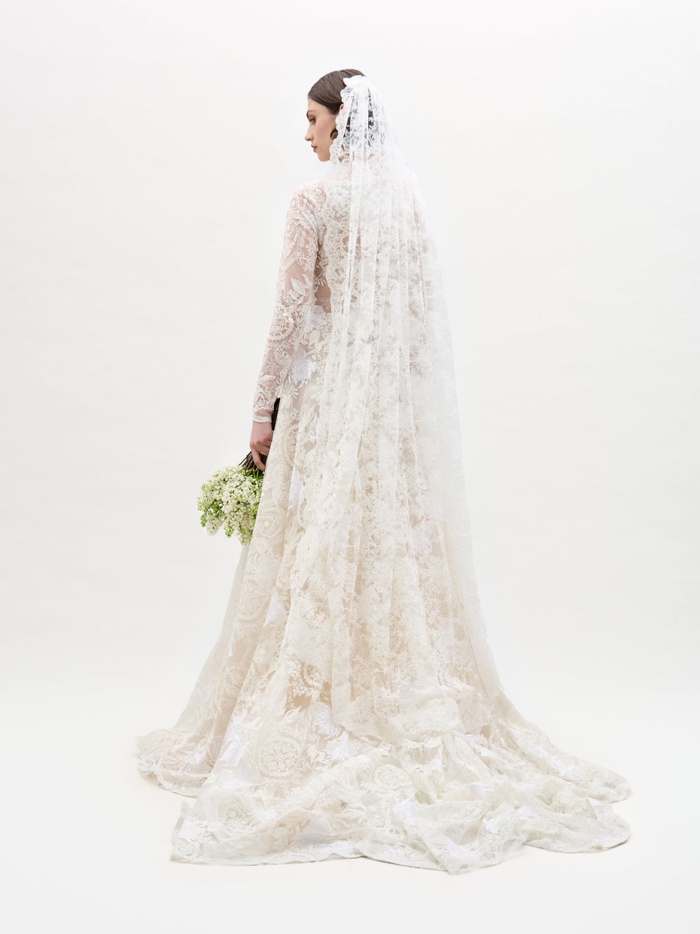 A bride, facing away from the camera, wears the Hermione Dress by SVT DESIGNS—a long lace wedding dress with a veil. She holds a small bouquet of white flowers against a plain white backdrop.