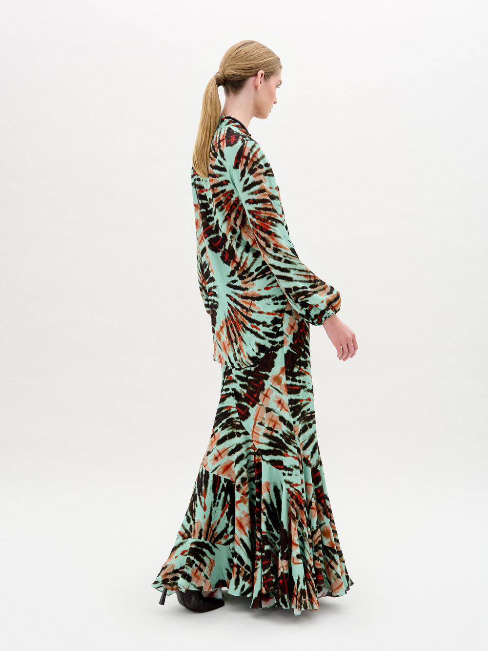 Wearing the SVT DESIGNS Florina Skirt, a person with a ponytail and long hair is seen in a tie-dye patterned dress featuring green, orange, and black shades. The individual, in a mermaid silhouette ensemble, is slightly turned to the left while walking on a plain white background.