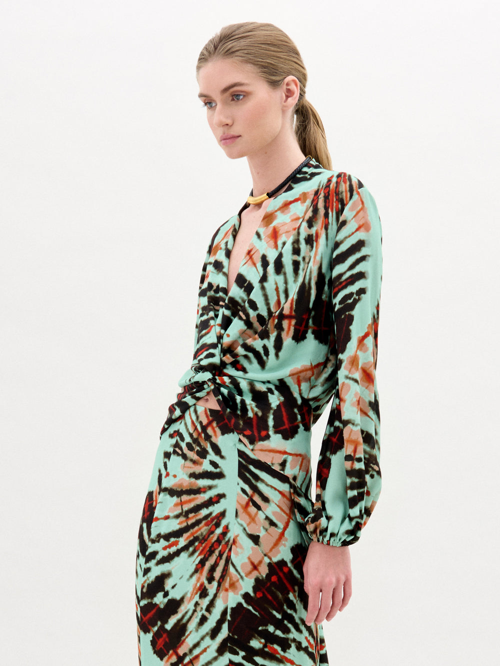 A woman with blonde hair in a low ponytail wears the SVT DESIGNS Honey Blouse, crafted from luxurious Italian fabric with a turquoise, black, and rust abstract pattern, standing against a plain white background.