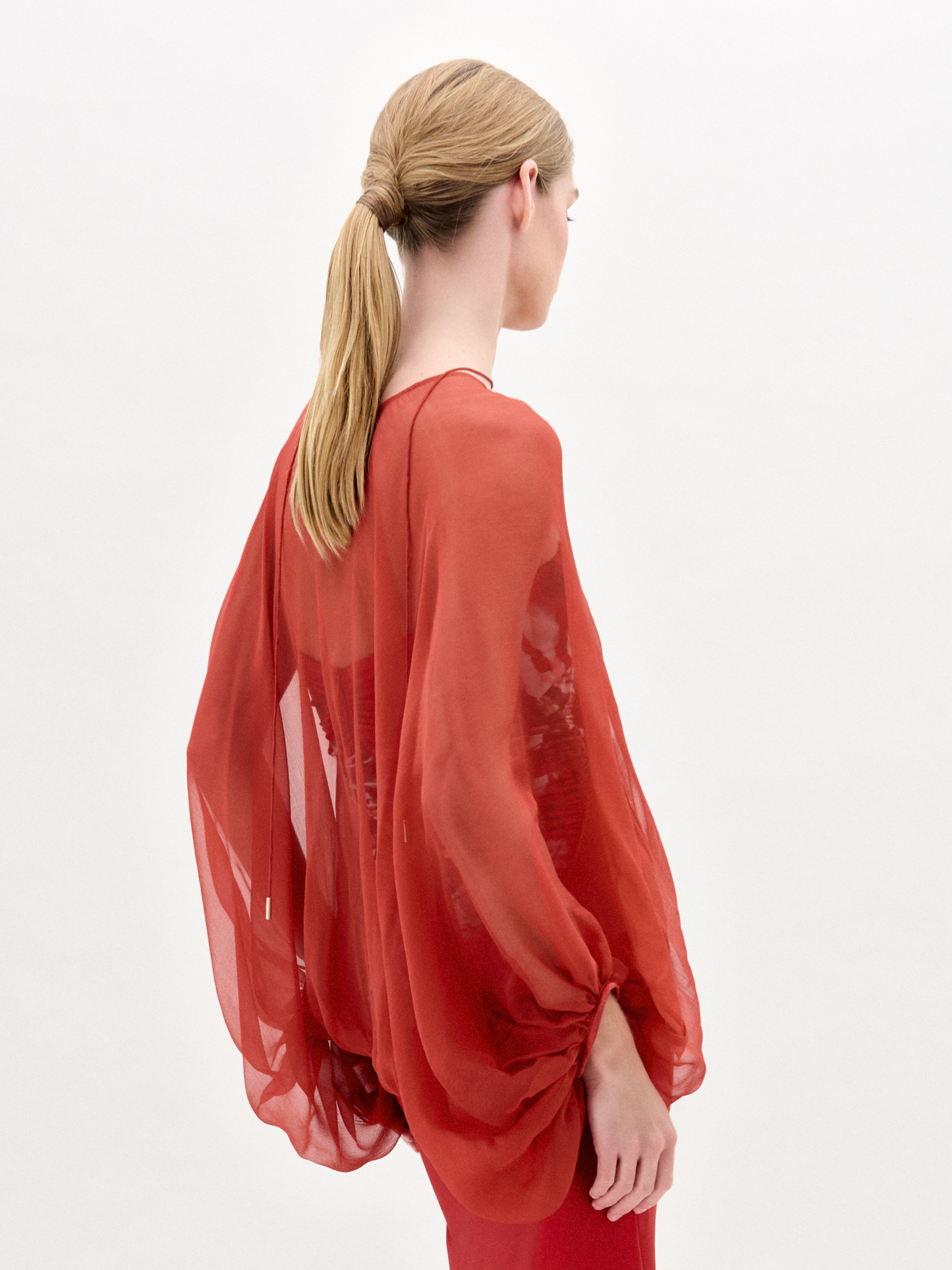 A woman stands against a plain background wearing the Idra Blouse, a sheer, long-sleeved top with a deep V neckline, layered over a patterned shirt and paired with a matching red skirt.