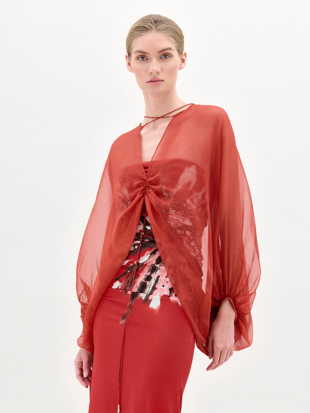 A person with light hair pulled back wears the Idra Blouse by SVT DESIGNS—a sheer red silk top with a deep V neckline—layered over a red and black patterned garment and a red skirt, posed against a plain white background.