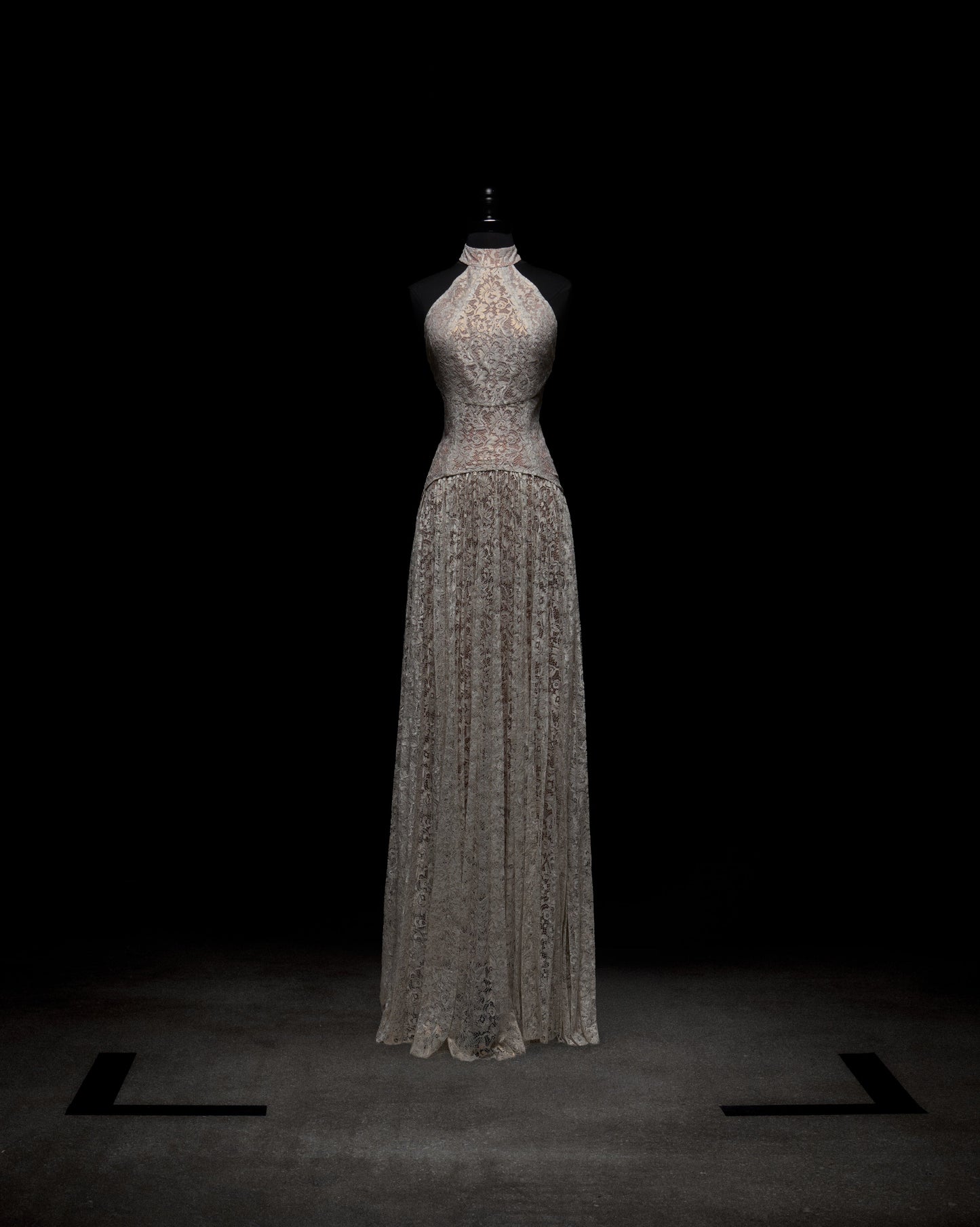 The Ilana Dress by SVT DESIGNS is a sleeveless, floor-length evening gown with a high neck, showcased on a mannequin against a dark background. Crafted from delicate lace with intricate patterns, it exudes elegance.