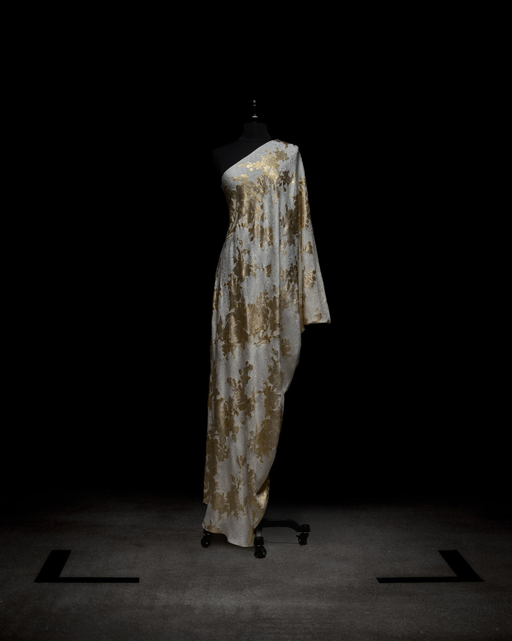 Displayed against a dark backdrop, the Inu Dress by SVT DESIGNS features a light fabric with shimmering gold floral patterns. This one-shoulder gown elegantly drapes to the floor with one sleeve over an arm; showcased on a black mannequin.