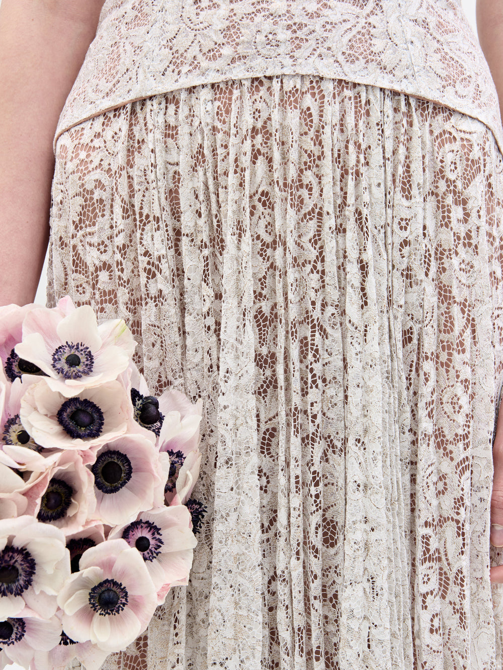 A close-up features someone in the Ilana Dress by SVT DESIGNS, holding a bouquet of light pink anemones with dark centers. The intricate floral lace pattern contrasts beautifully with the delicate blooms.