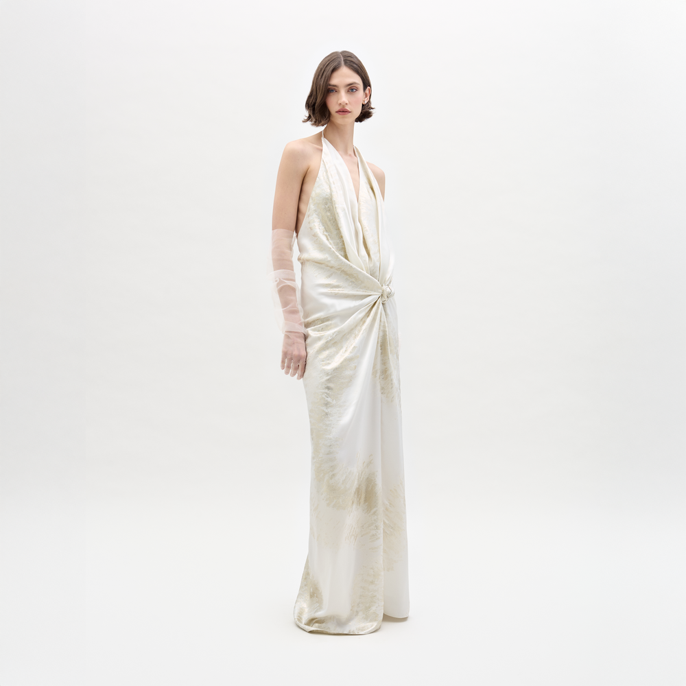 A woman stands against a white background wearing the Jazmin Dress by SVT DESIGNS, a long sleeveless maxi with a draped halter neckline, gathered waist knot, and one arm elegantly covered in sheer Italian fabric.