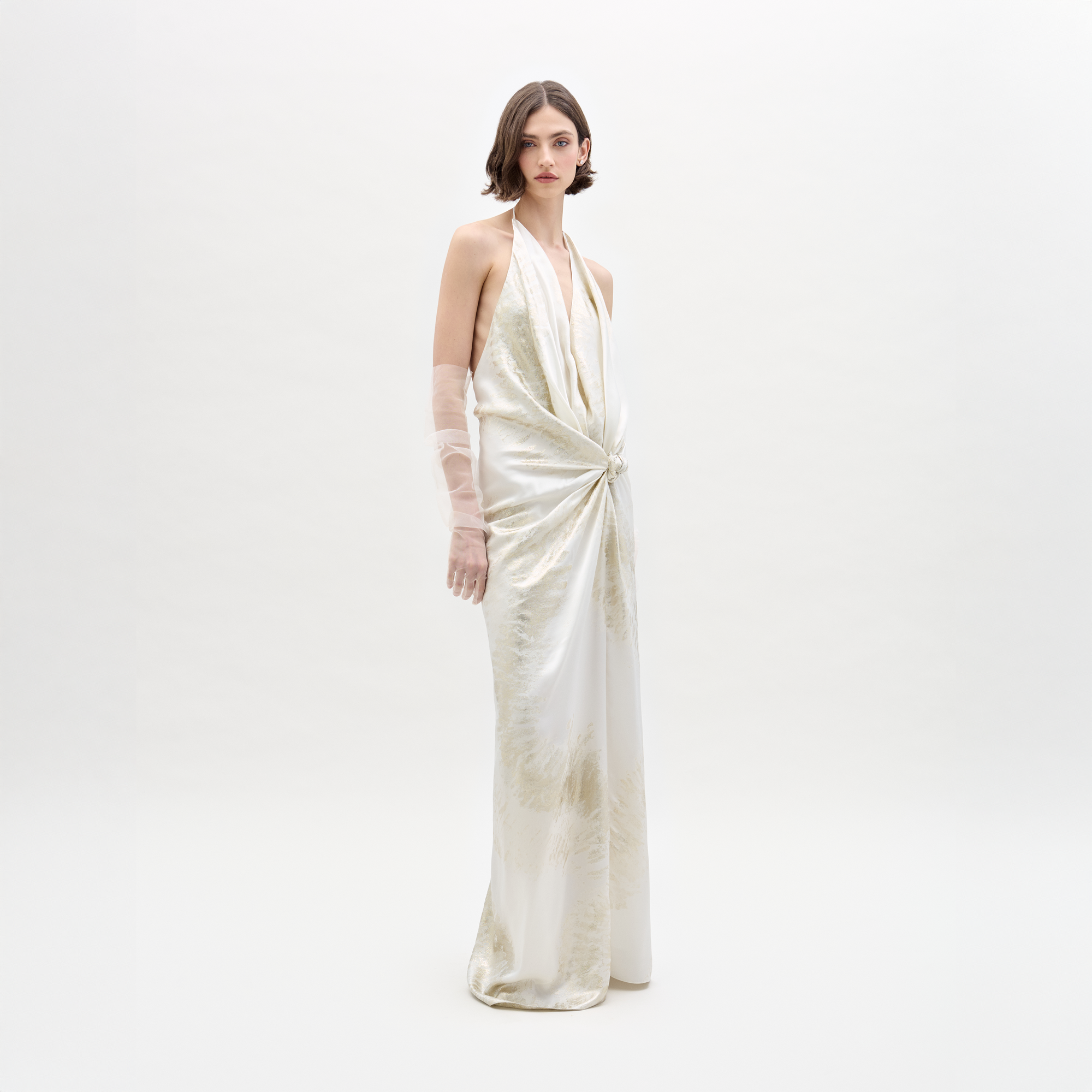 A woman stands against a white background wearing the Jazmin Dress by SVT DESIGNS, a long sleeveless maxi with a draped halter neckline, gathered waist knot, and one arm elegantly covered in sheer Italian fabric.
