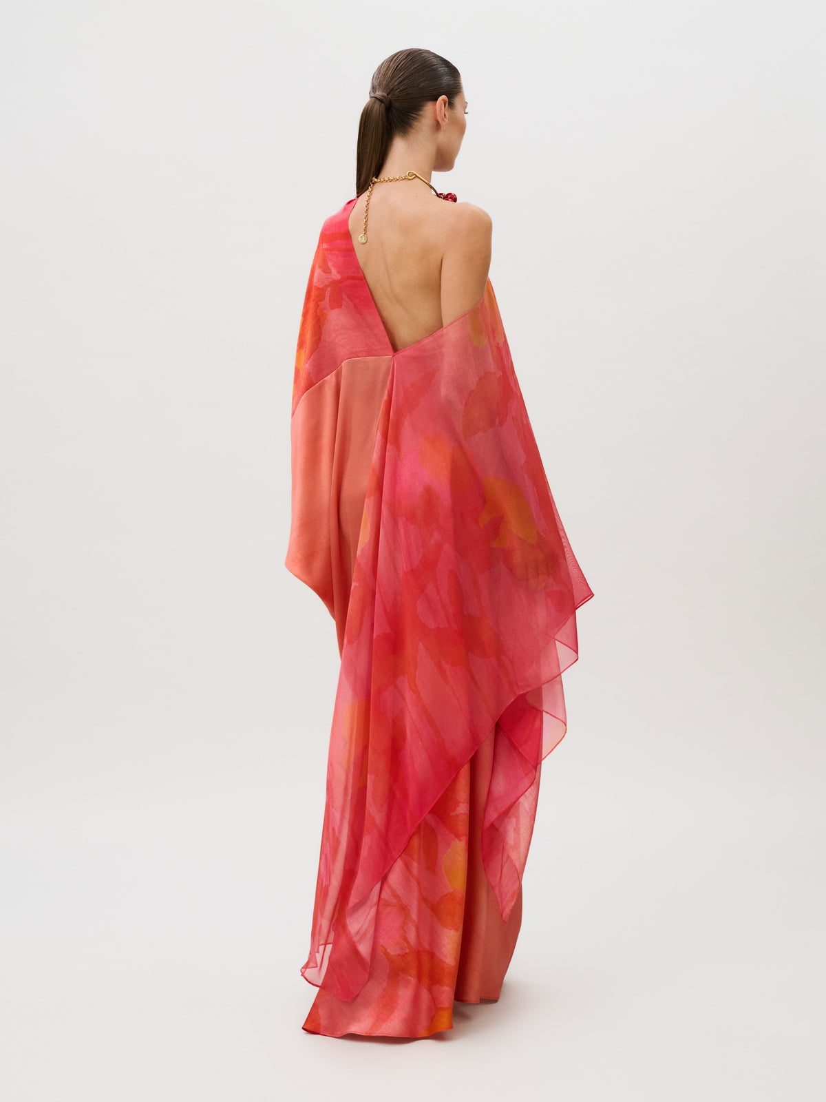A person viewed from behind wearing the Jade Dress, a sleeveless, floor-length Italian silk chiffon gown with an abstract pink and orange floral pattern and draped fabric panels.