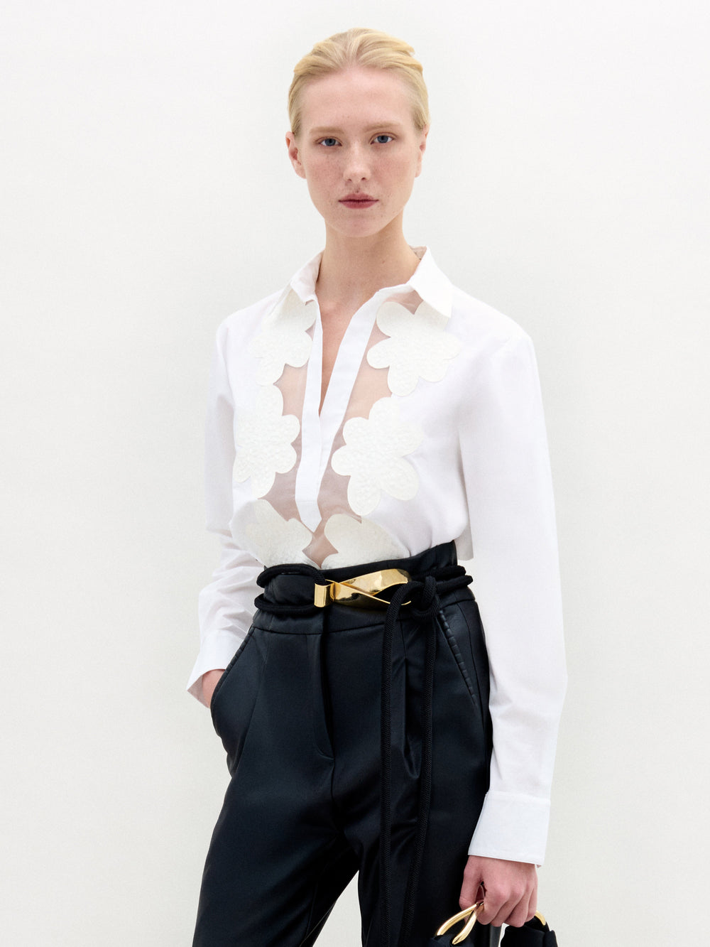 A person stands against a plain white background wearing the Karole Blouse by SVT DESIGNS, featuring a long-sleeve white design with sheer floral appliqué on the front, paired with black high-waisted pants and holding a pair of glasses.