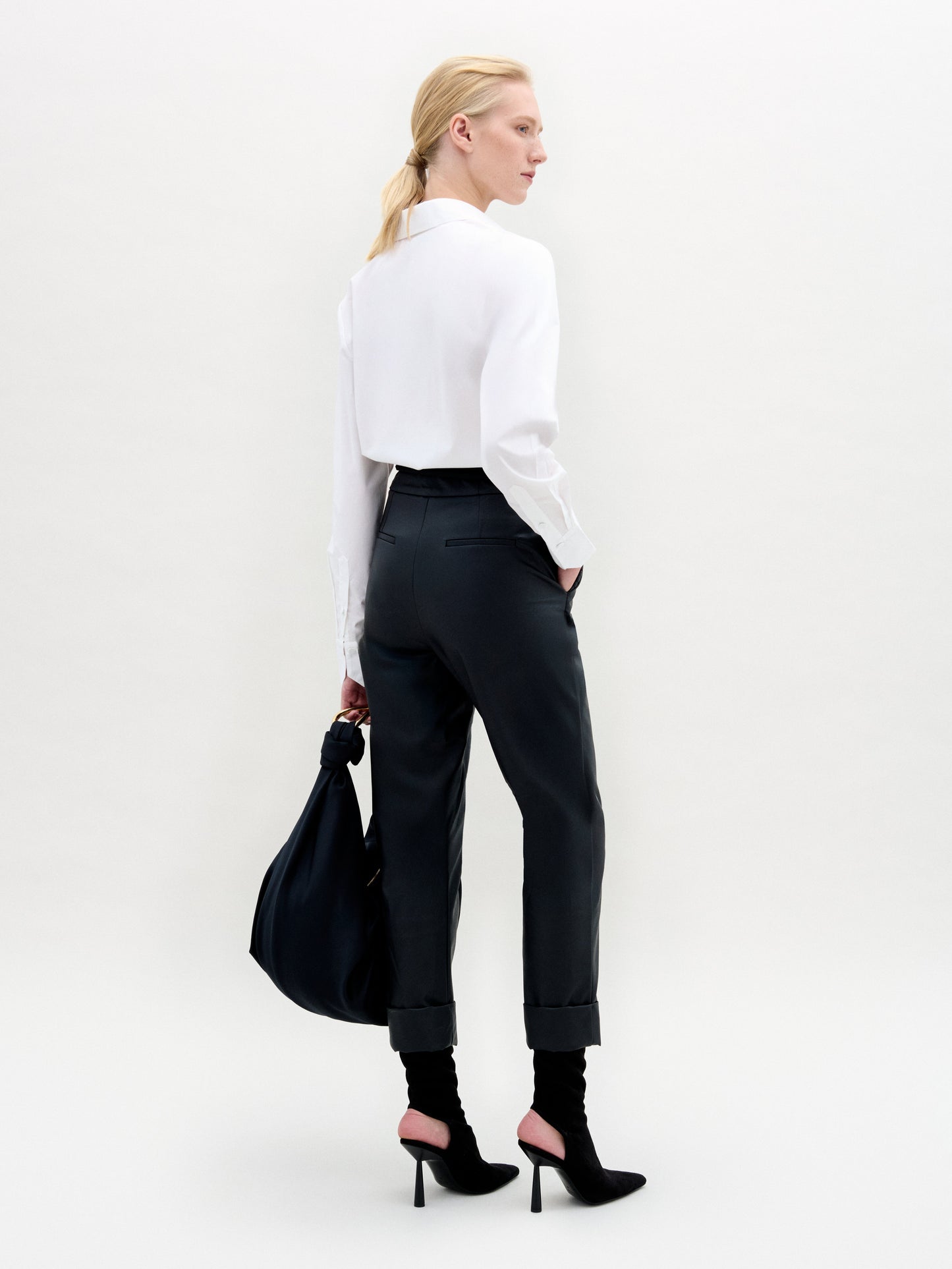Woman wearing a white blouse and black pants holding a black bag against a white background