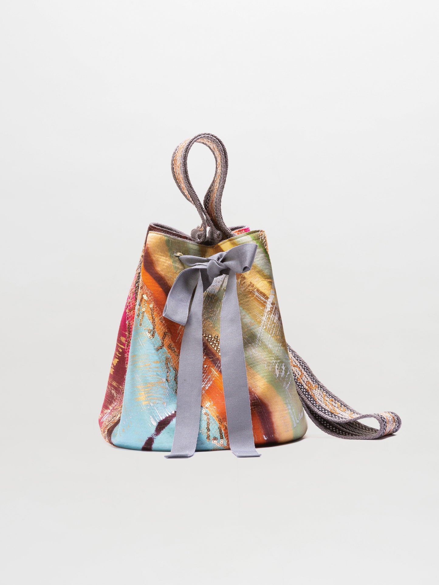 The SVT DESIGNS Mochila Laaput Small is a bucket-shaped handbag featuring a multicolored abstract pattern, a gray fabric bow on the front, and a long woven strap, shown against a plain light background.
