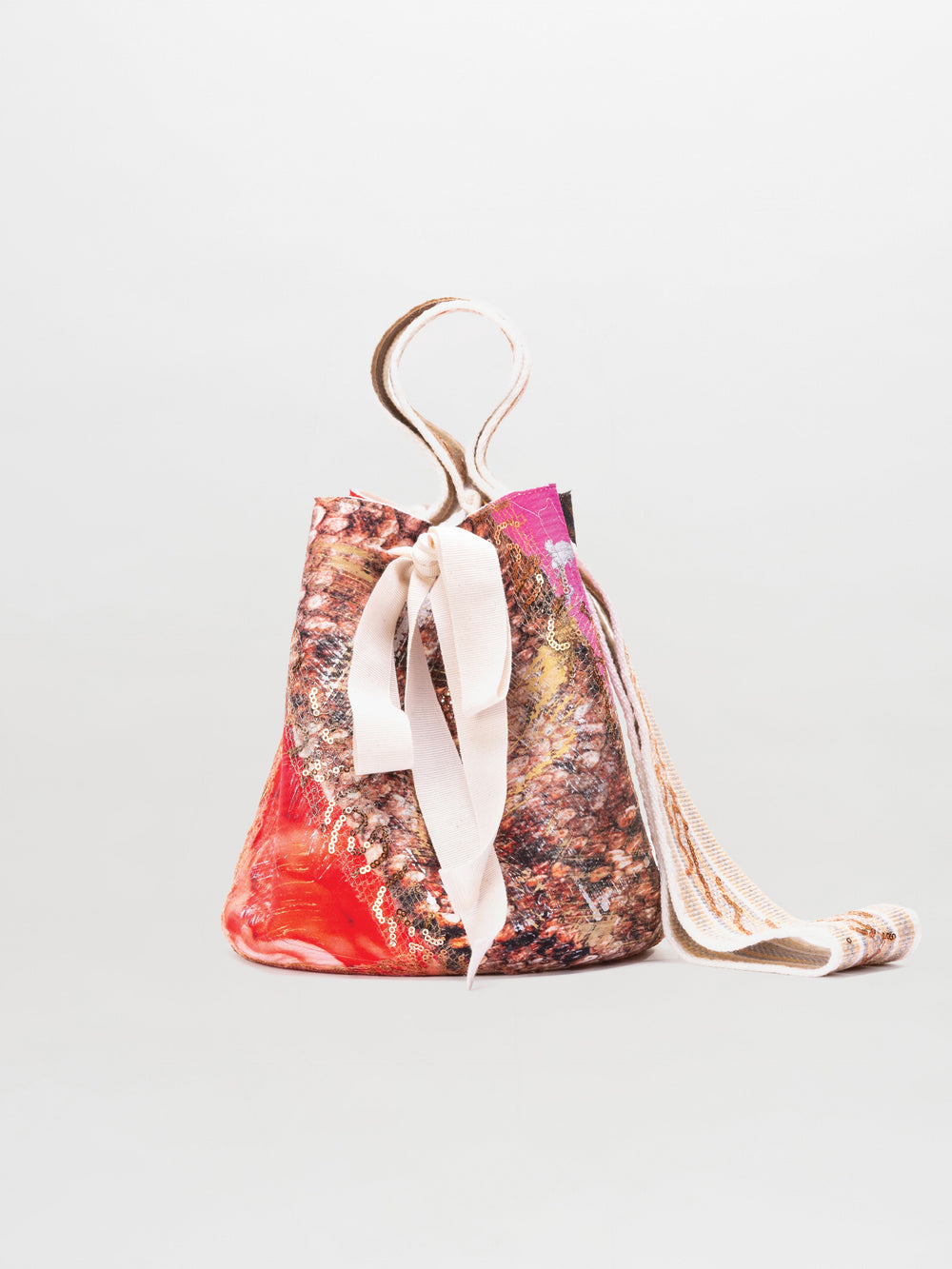 The SVT DESIGNS Mochila Laaput Small is a drawstring bucket bag inspired by Wayuu craftsmanship, featuring a multicolored pattern in pink, red, beige, and brown, a wide fabric strap, and a pale ribbon on a plain light gray background.