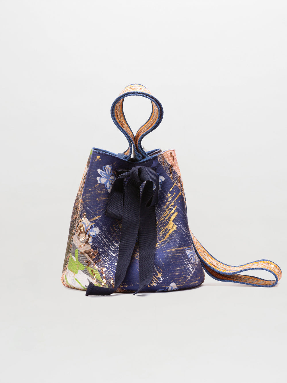The SVT DESIGNS Mochila Laaput Big is a bucket-style mochila handbag featuring multicolored patterned fabric, a dark front bow, and a wide hand-woven Wayuu-inspired strap, shown against a plain white background.