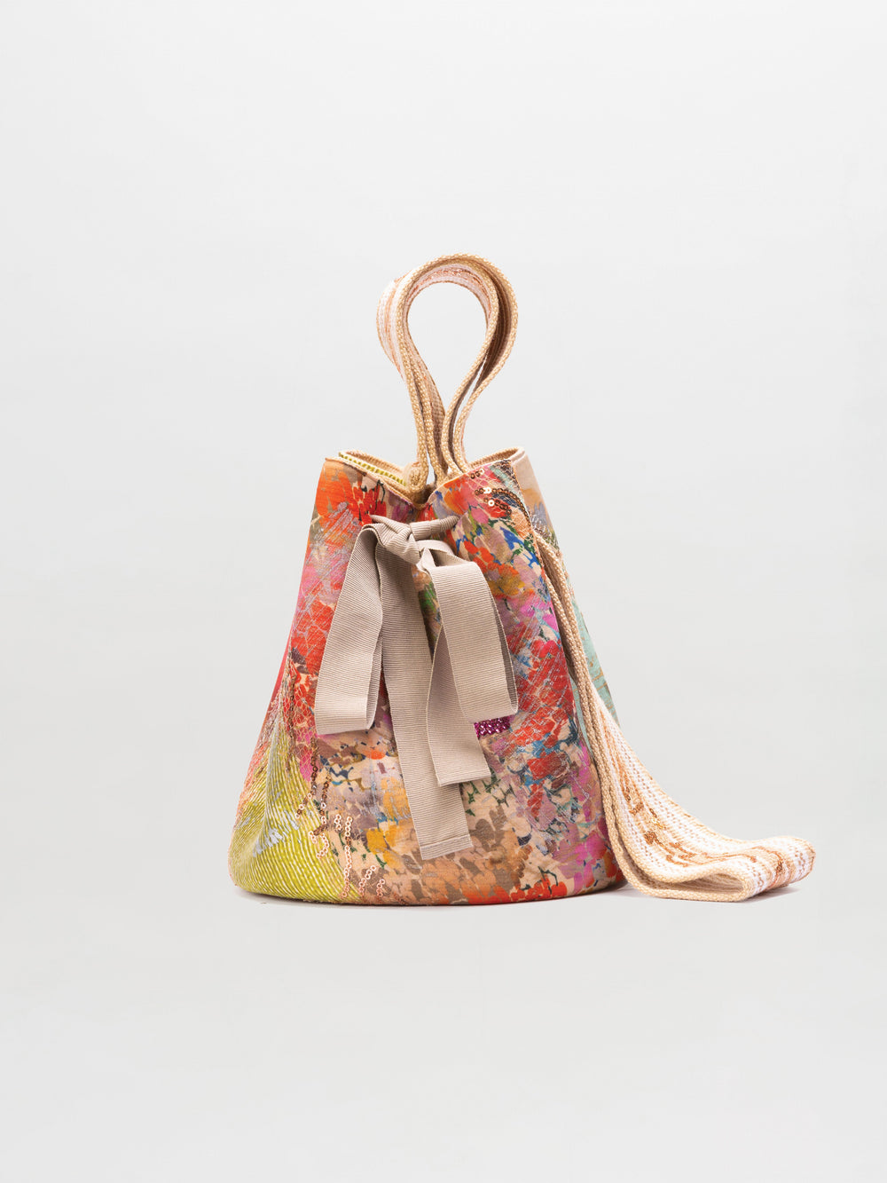 The Mochila Laaput Big by SVT DESIGNS is a multicolored Wayuu handbag with a floral pattern, beige fabric handles, and a beige ribbon bow on the front, beautifully set against a plain light gray background.