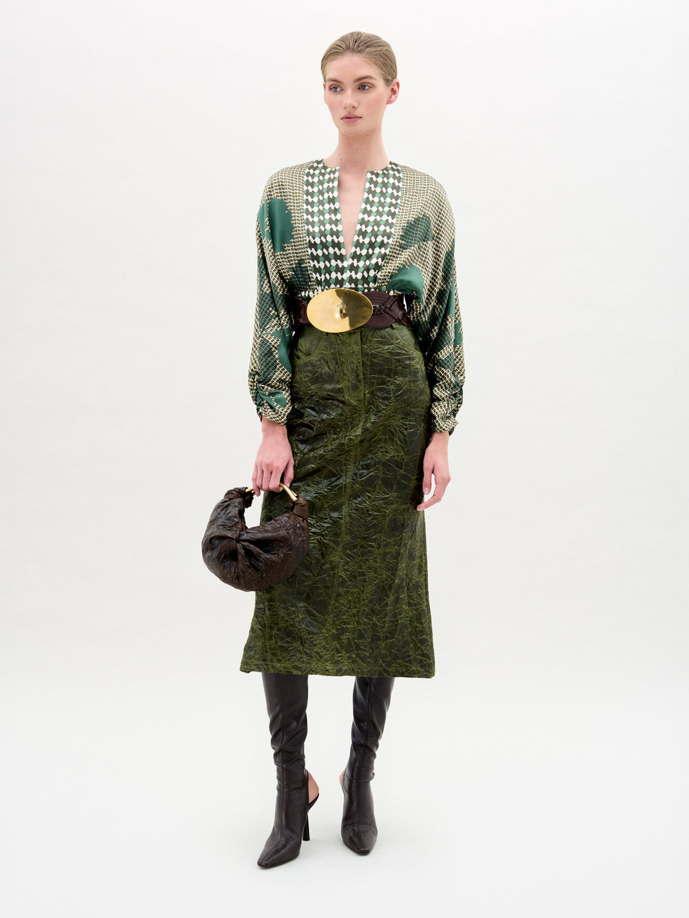 A woman stands against a plain background wearing a patterned green blouse, a wide belt with a large gold buckle, SVT DESIGNS' Layan Skirt made in Colombia, black knee-high boots, and holding a brown handbag.