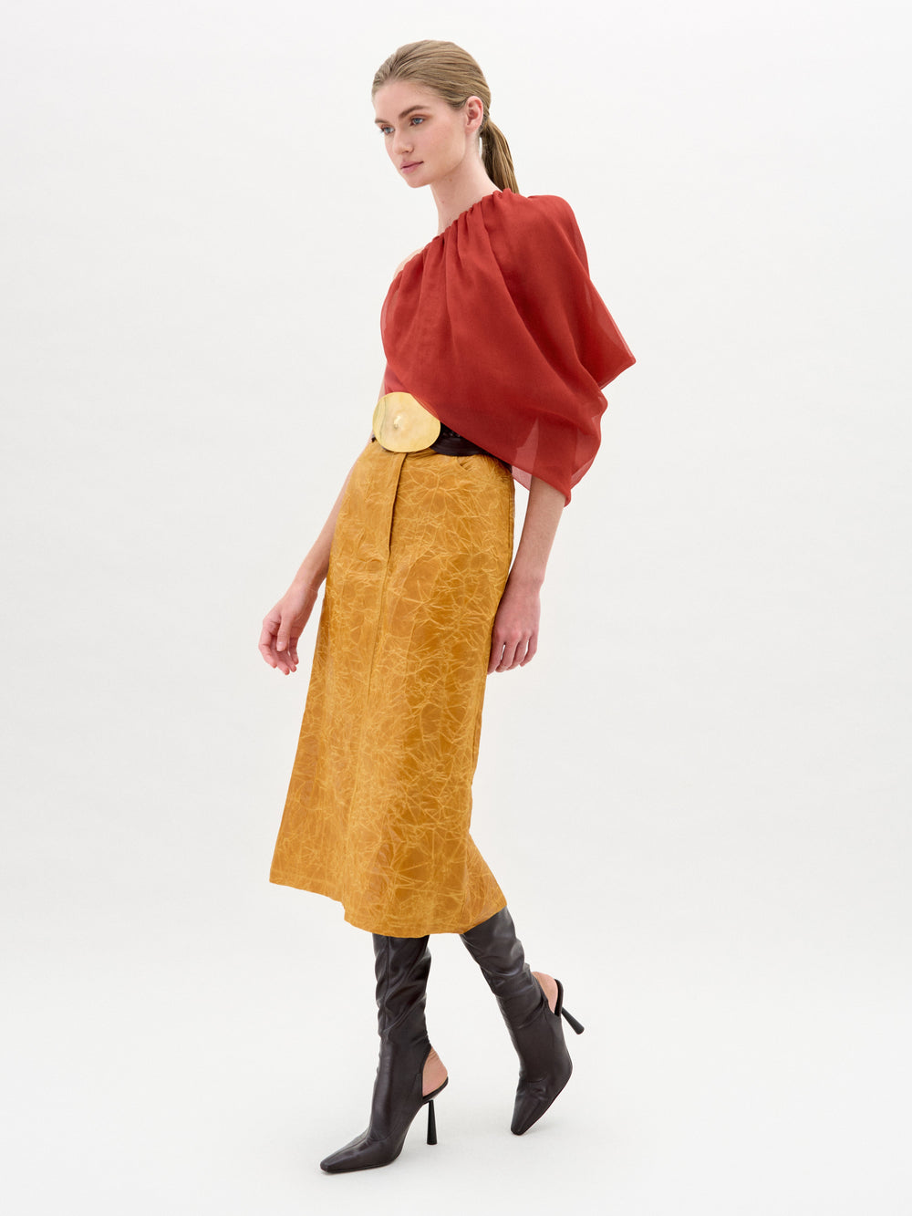 A woman stands against a white background wearing a sheer red blouse, the Layan Skirt by SVT DESIGNS in textured mustard yellow Italian fabric, a wide belt with a large buckle, and knee-high dark brown heeled boots.