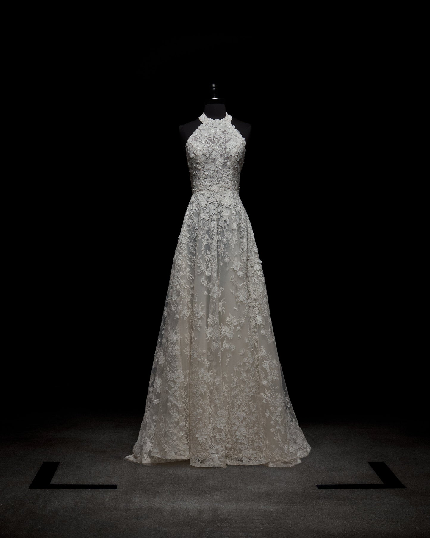The Leia Dress by SVT DESIGNS, a white lace halter-neck piece with intricate floral patterns, a fitted bodice, and a flowing skirt, is showcased on a black mannequin against a dark background.