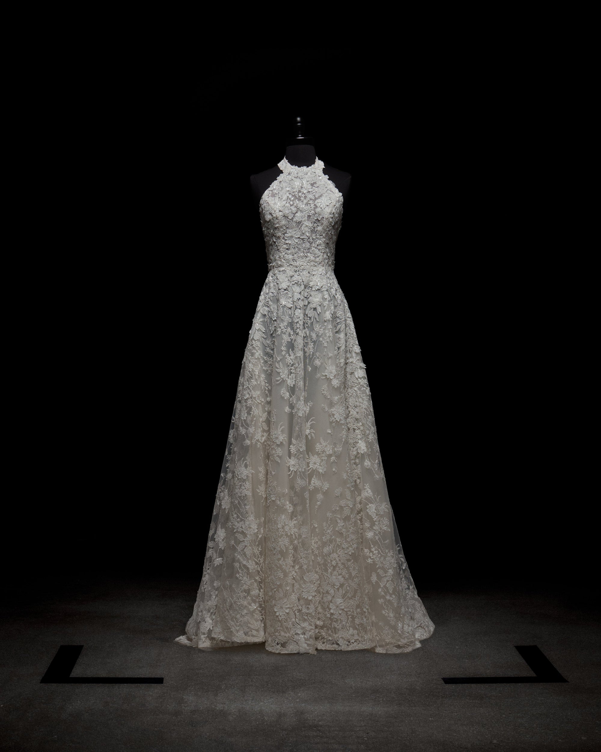 The Leia Dress by SVT DESIGNS, a white lace halter-neck piece with intricate floral patterns, a fitted bodice, and a flowing skirt, is showcased on a black mannequin against a dark background.