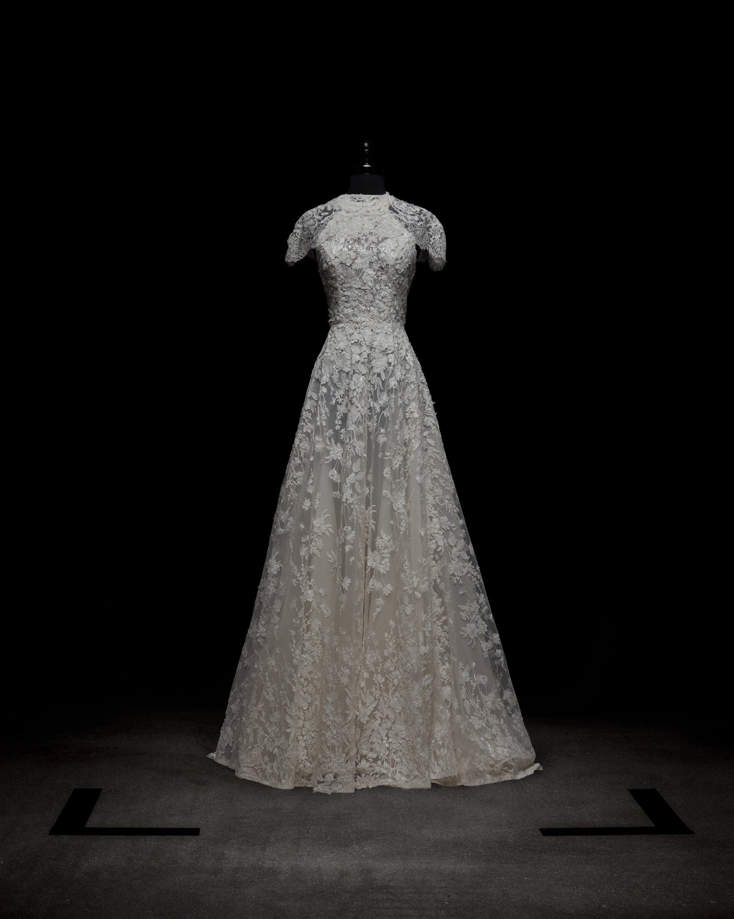 The Leia Dress with Cape by SVT DESIGNS, a white lace floor-length gown with short sleeves and a fitted bodice, is showcased on a black mannequin against a dark background. Black markers form a square on the floor around the dress.