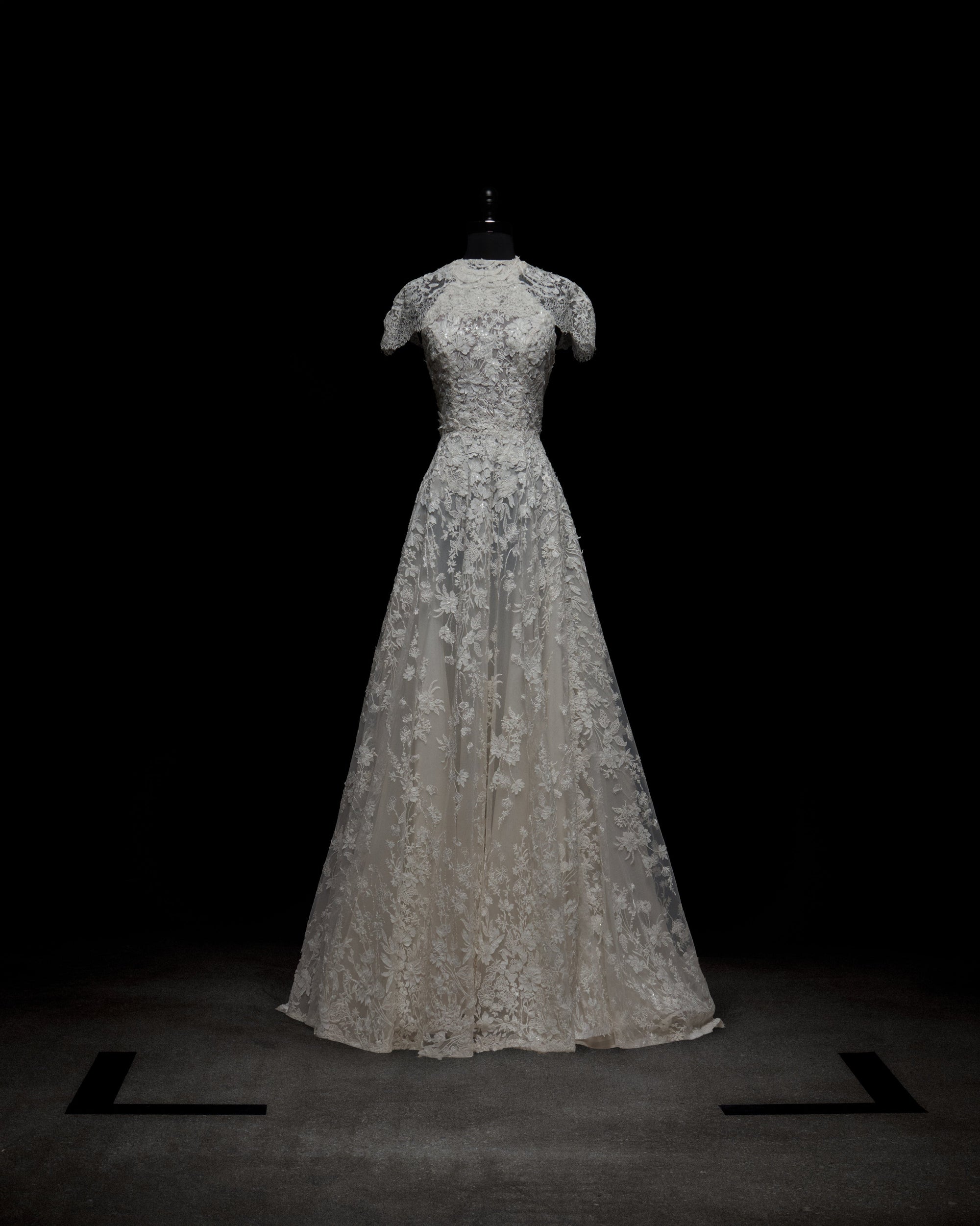 The Leia Dress with Cape by SVT DESIGNS, a white lace floor-length gown with short sleeves and a fitted bodice, is showcased on a black mannequin against a dark background. Black markers form a square on the floor around the dress.