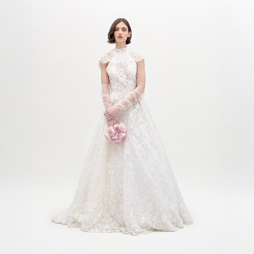 A woman wears the SVT DESIGNS Leia Dress with Cape, a full-length white lace wedding gown with delicate detailing and sheer gloves, holding light pink flowers against a plain white background.
