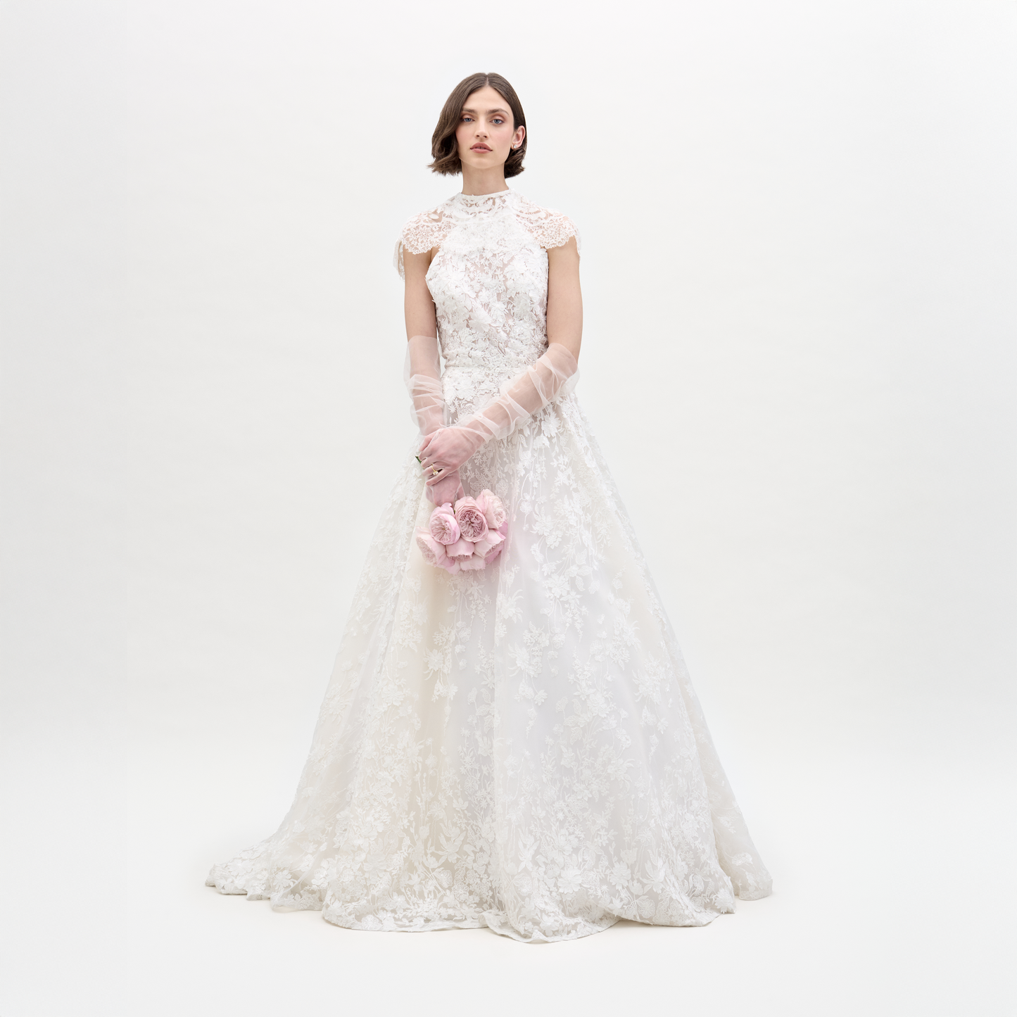 A woman wears the SVT DESIGNS Leia Dress with Cape, a full-length white lace wedding gown with delicate detailing and sheer gloves, holding light pink flowers against a plain white background.