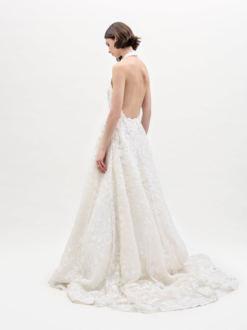 A woman with short brown hair stands on a white background wearing SVT DESIGNS' Leia Dress, a white, backless lace gown featuring a halter neck and long train. She faces sideways with her head slightly turned down.