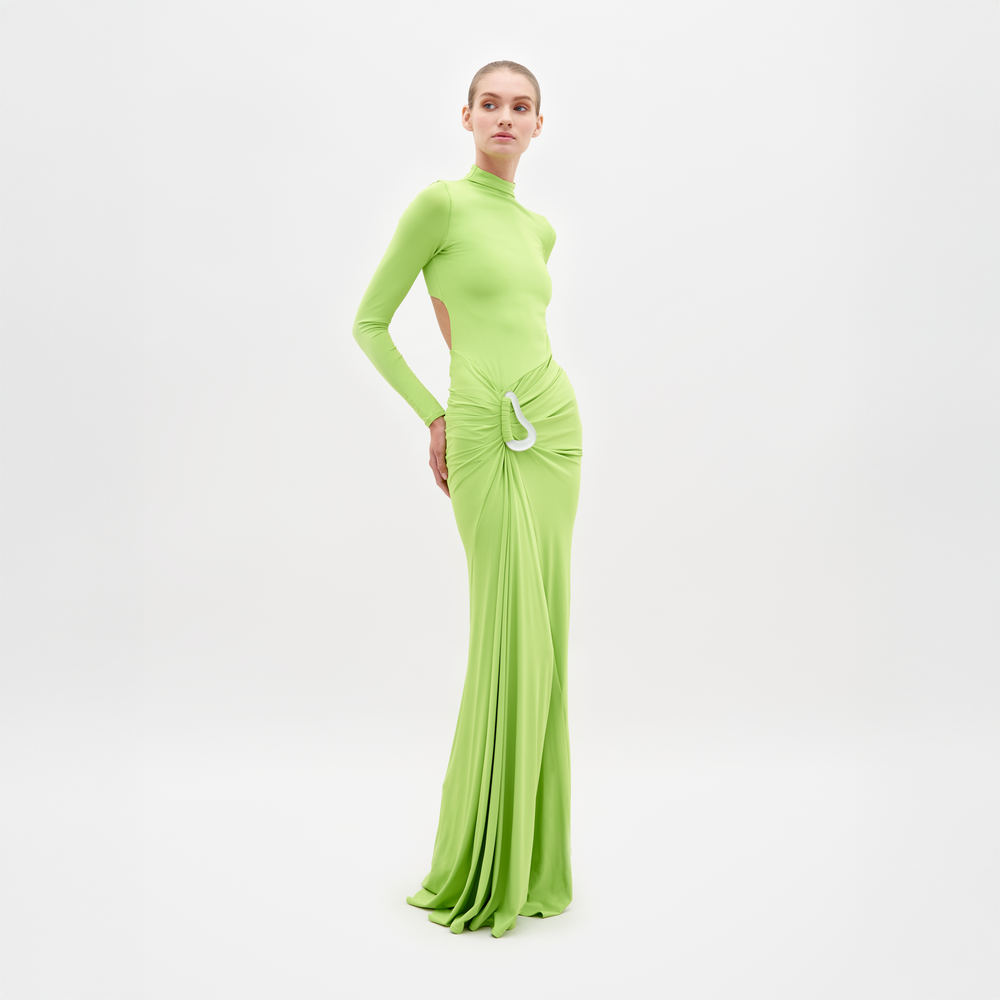 A person stands wearing SVT DESIGNS' Lena Dress, a floor-length maxi in bright green with cut-out sides and gathered waist detail, made from fine Italian textile. The simple white backdrop enhances its luxurious look.