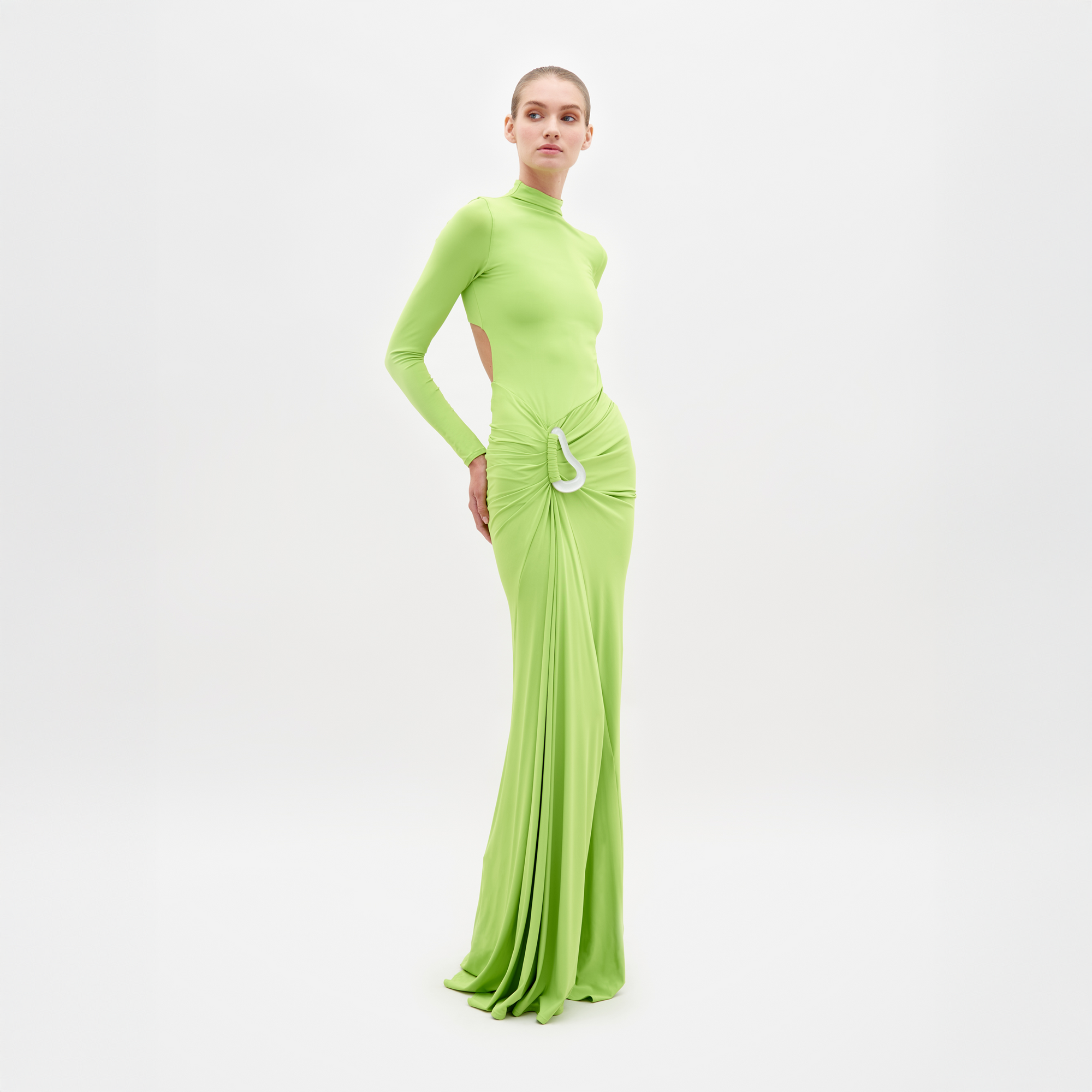 A person stands wearing SVT DESIGNS' Lena Dress, a floor-length maxi in bright green with cut-out sides and gathered waist detail, made from fine Italian textile. The simple white backdrop enhances its luxurious look.