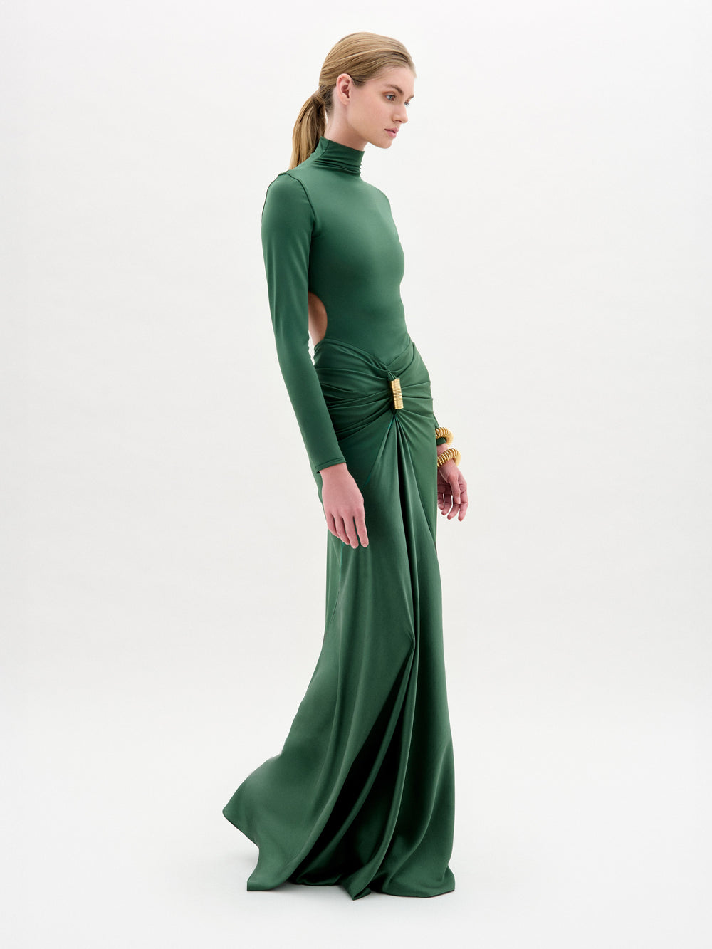 A woman with blonde hair pulled back models the SVT DESIGNS Lena Dress—a fitted, long-sleeved green maxi with side cutouts and draped details—against a plain white background, accessorized with gold bracelets.
