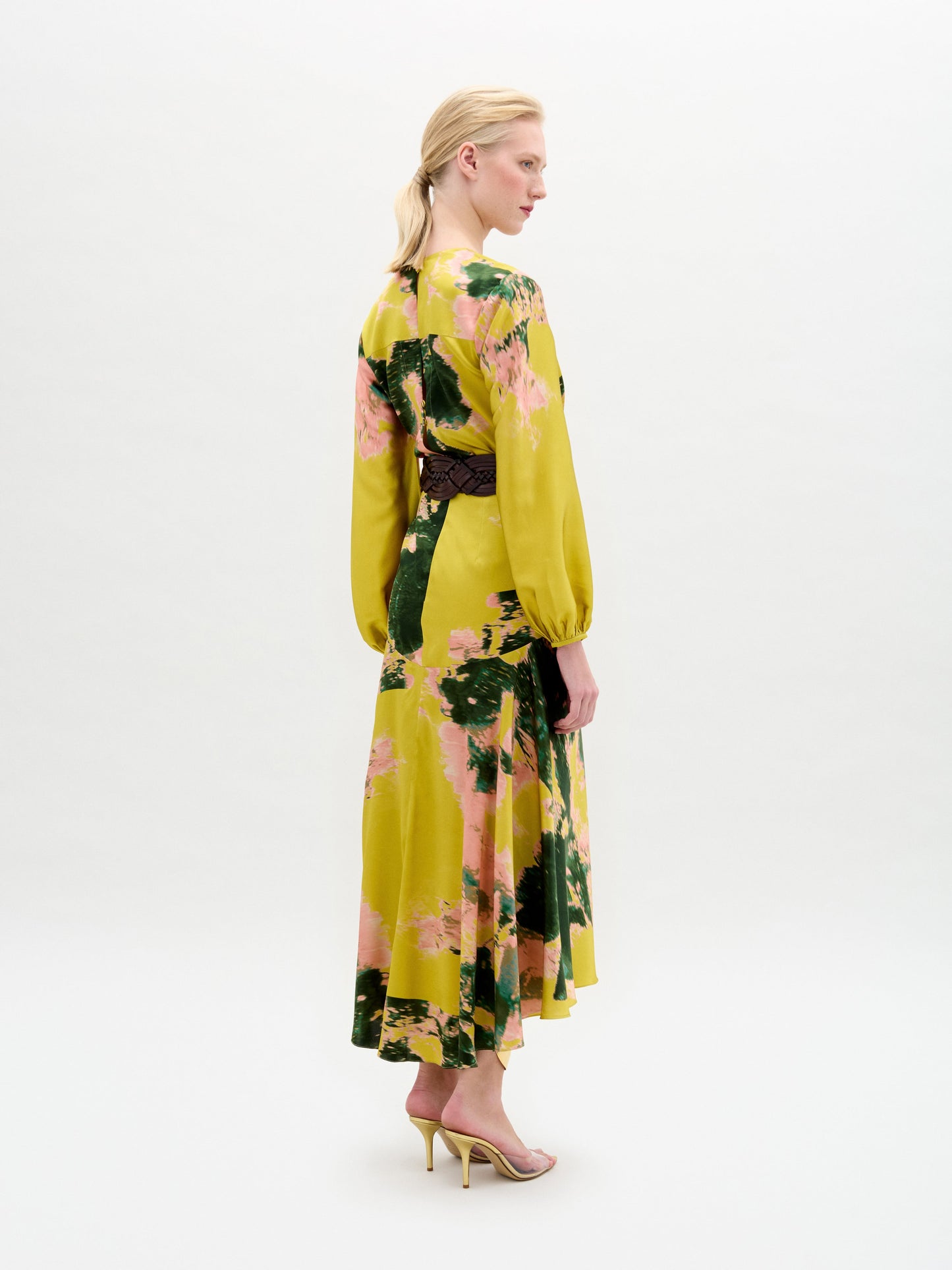 A woman in profile wears the Lima Dress by SVT DESIGNS, an Italian-fabric maxi with pink and green florals, a dark belt, and yellow heels, shown against a plain white background.