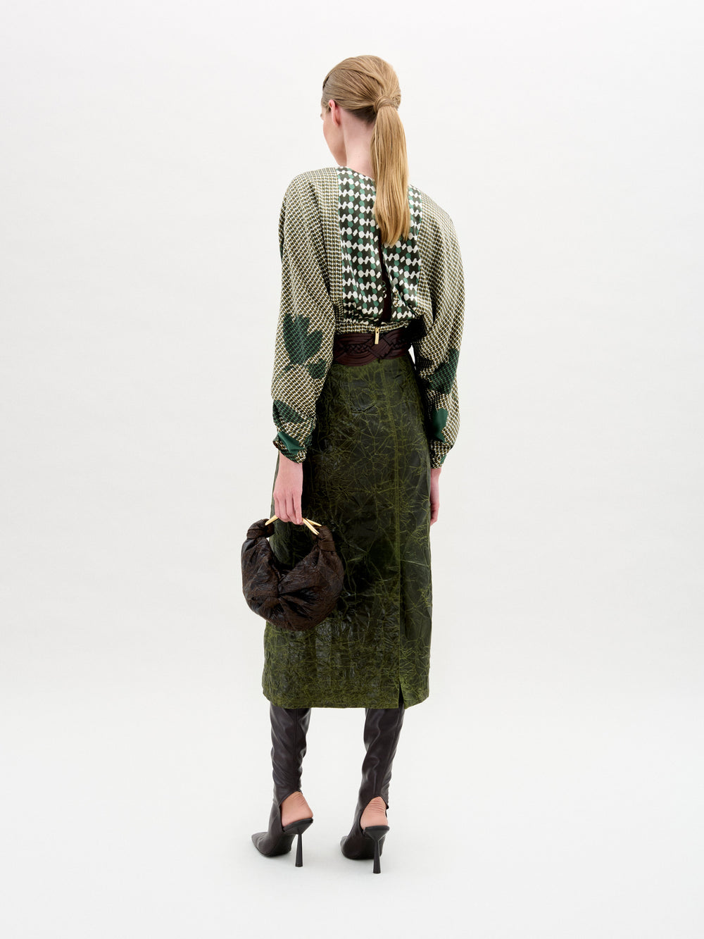 A person with long blond hair tied back faces away, wearing a green patterned top, the Layan Skirt by SVT DESIGNS (dark green, structured, made in Colombia), a brown belt, dark brown heeled boots, and holding a dark brown bag against a plain white background.