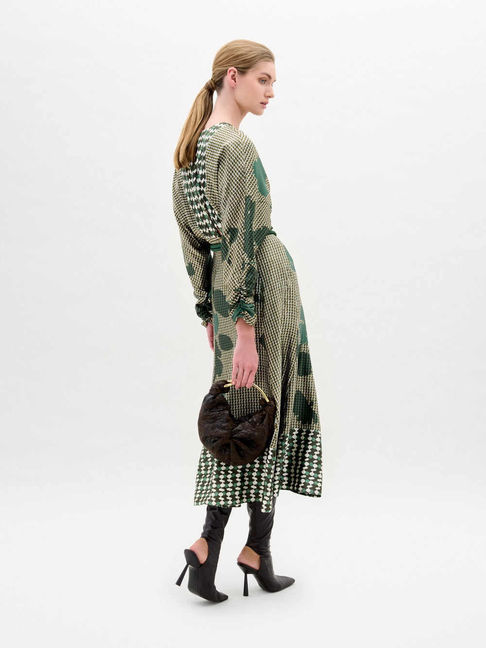A blonde-haired person in a ponytail wears the SVT DESIGNS Erin Skirt, featuring a green and cream pattern, paired with black heeled boots and holding a round dark brown handbag against a plain white background.