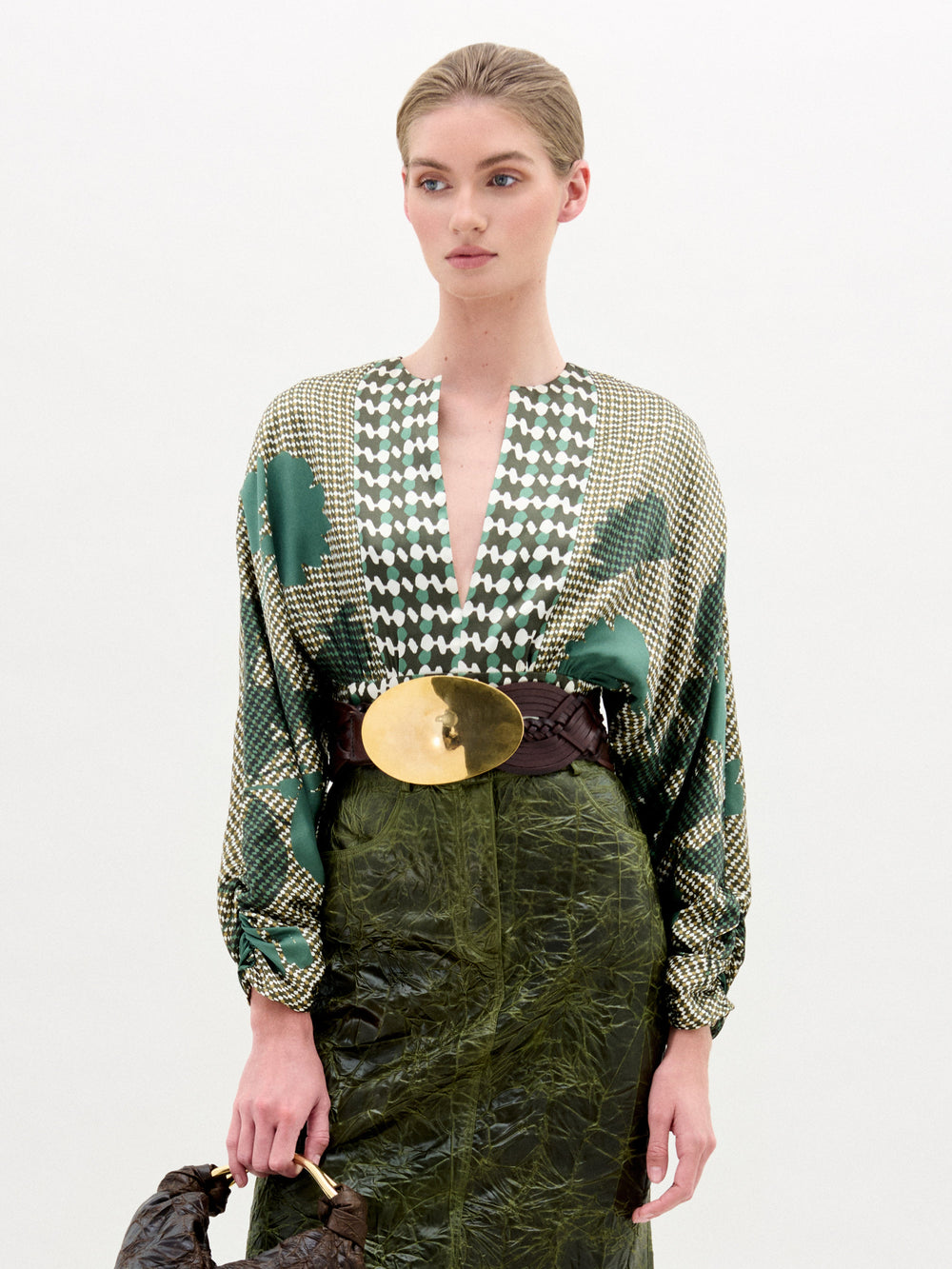 A woman stands against a plain background wearing the Lizzy Blouse by SVT DESIGNS, a patterned silk top with a deep V-neck. She pairs it with a large gold belt, textured green skirt, brown handbag, and has her hair pulled back.