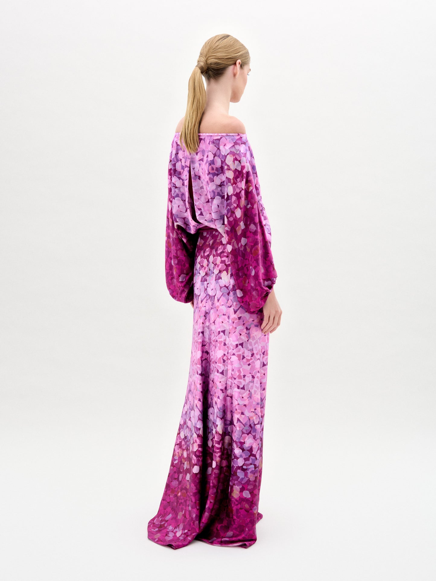 A person with long hair tied back is facing away, wearing the elegant Lorella Dress by SVT DESIGNS. This off-the-shoulder maxi dress features flowing purple and pink Italian fabric adorned with a floral pattern, set against a plain, light-colored background.