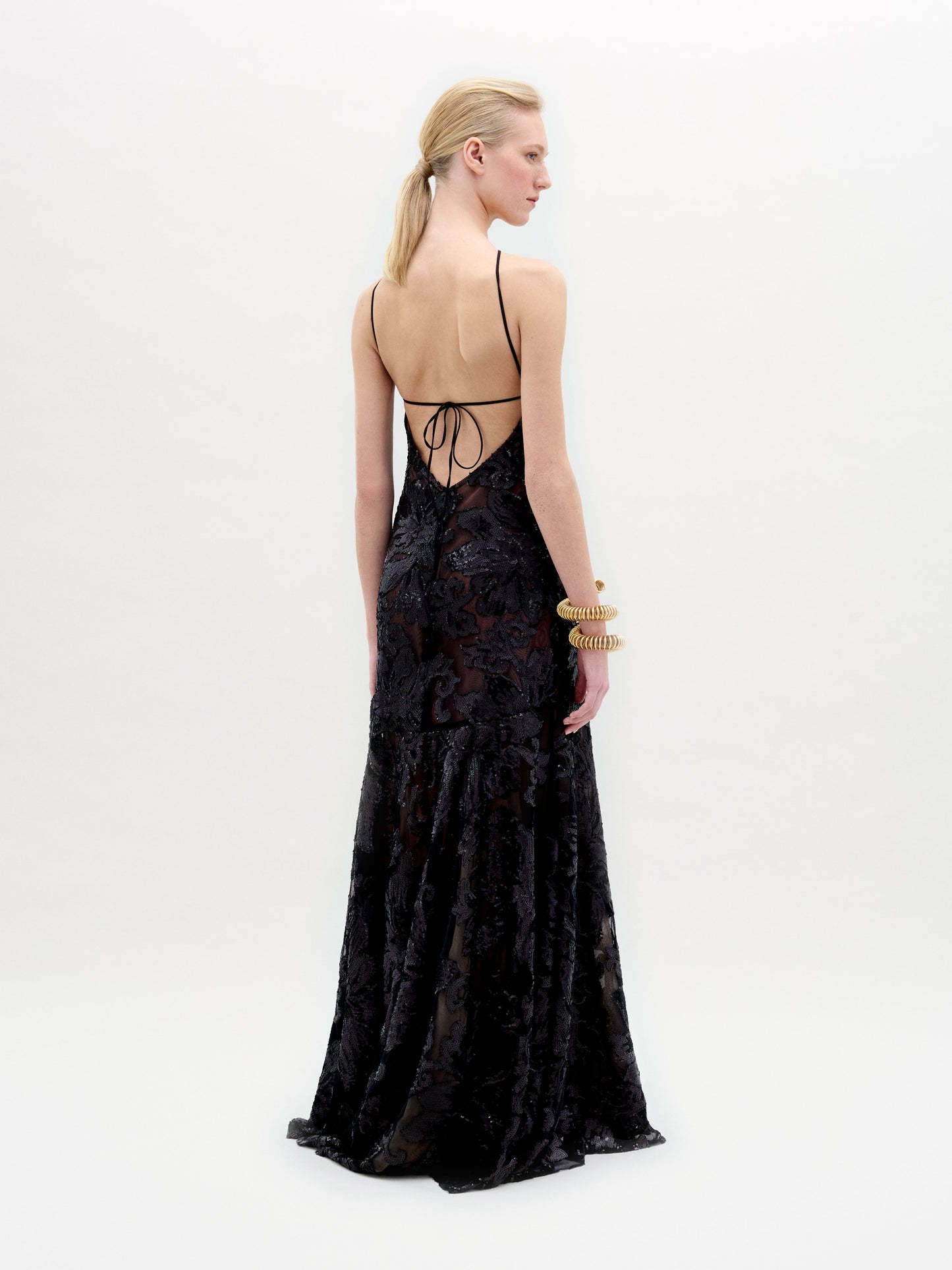 A woman with blonde hair tied back models the Lottise Dress—a long, black, floral-patterned dress made from Italian fabric with thin straps and a low, strappy back—paired with multiple gold bangles on her left arm.