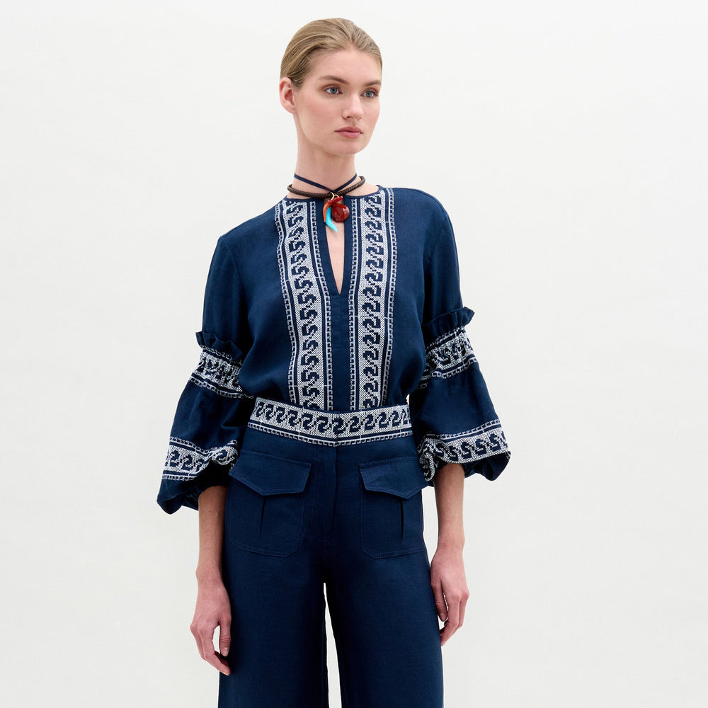 A person stands against a plain light background, wearing the Lucaya Blouse by SVT DESIGNS with voluminous sleeves and white embroidery, paired with matching high-waisted pants and a choker necklace featuring a red pendant.