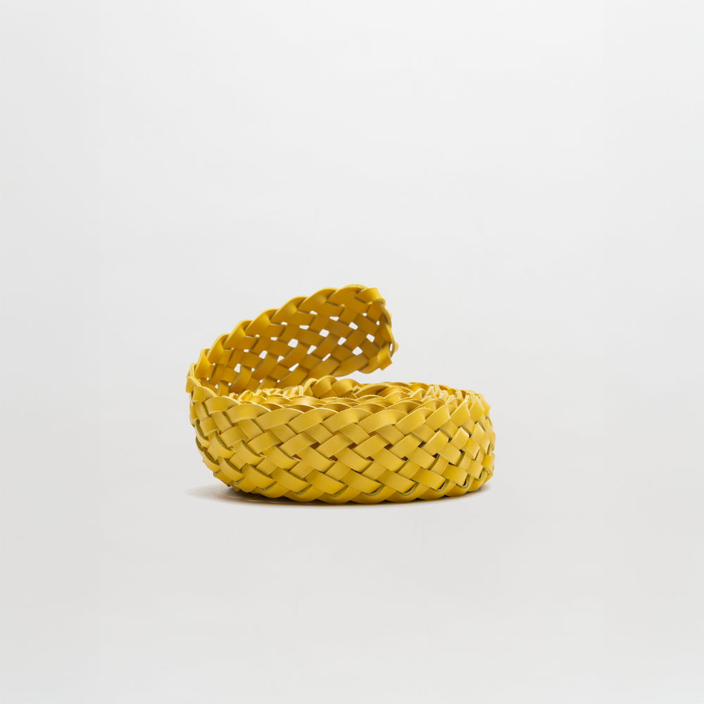 The Lauren Belt by SVT DESIGNS, a yellow braided leather accessory, is coiled on a white background with one end slightly raised to highlight its textured weave.