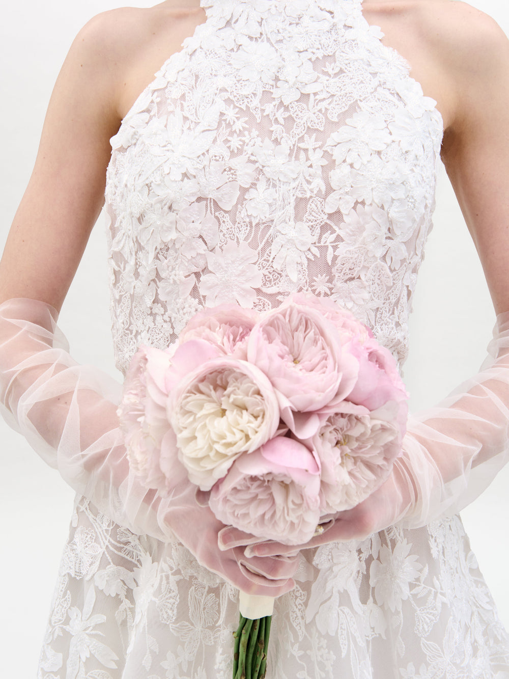 A bride wearing the Leia Dress by SVT DESIGNS, featuring a white lace design and sheer gloves, holds a bouquet of pale pink and white peonies against a plain white background.