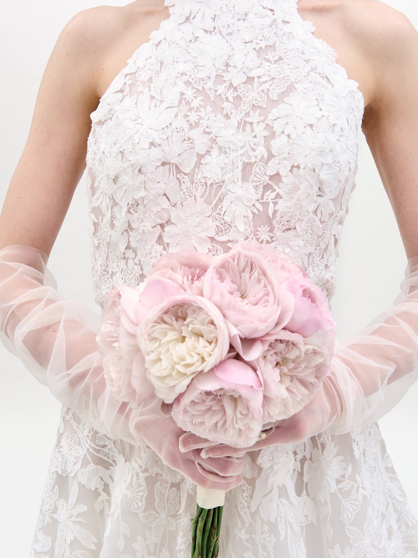 A bride wearing the Leia Dress by SVT DESIGNS, featuring a white lace design and sheer gloves, holds a bouquet of pale pink and white peonies against a plain white background.