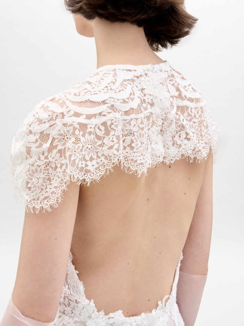 A person wears the Leia Dress with Cape by SVT DESIGNS, an intricate white lace dress with an open back, delicate floral patterns, and scalloped edges on the shoulders. The background is plain and light-colored.