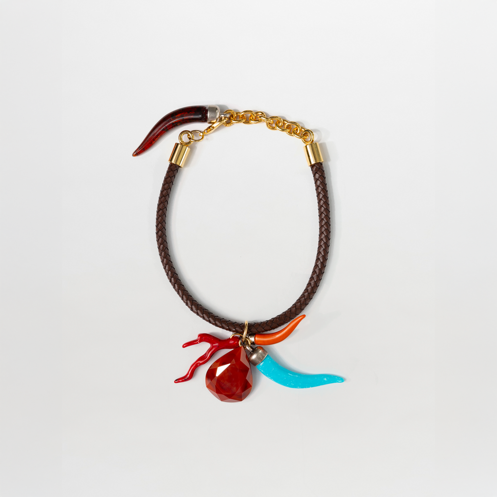 The Leslie Chocker by SVT DESIGNS features a brown braided band, gold-tone clasp and chain, and three multicolor charms: a red faceted gem, curved turquoise charm, and curved red charm with metallic accents.