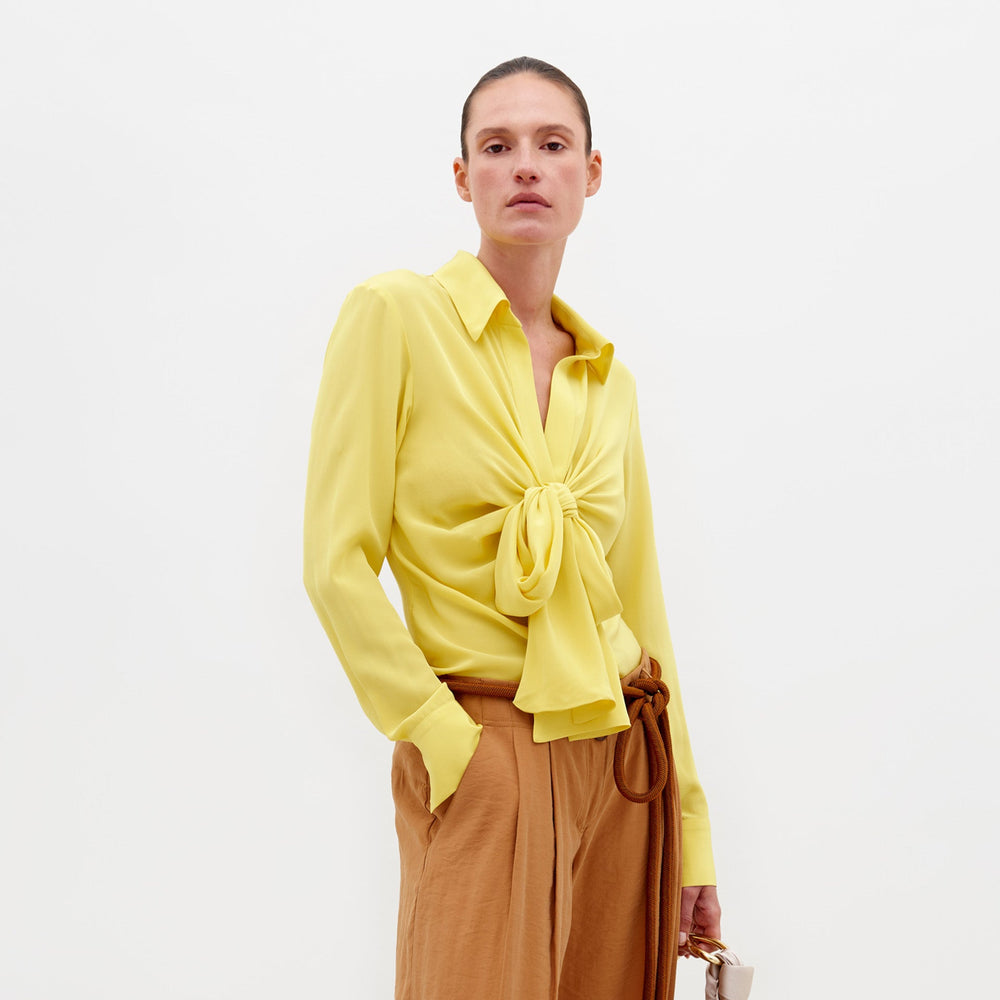 A person stands against a white background wearing the SVT DESIGNS Mercury Blouse Yellow with a front bow and classic collar, paired with light brown high-waisted pants. One hand is in their pocket; the other holds a small object.