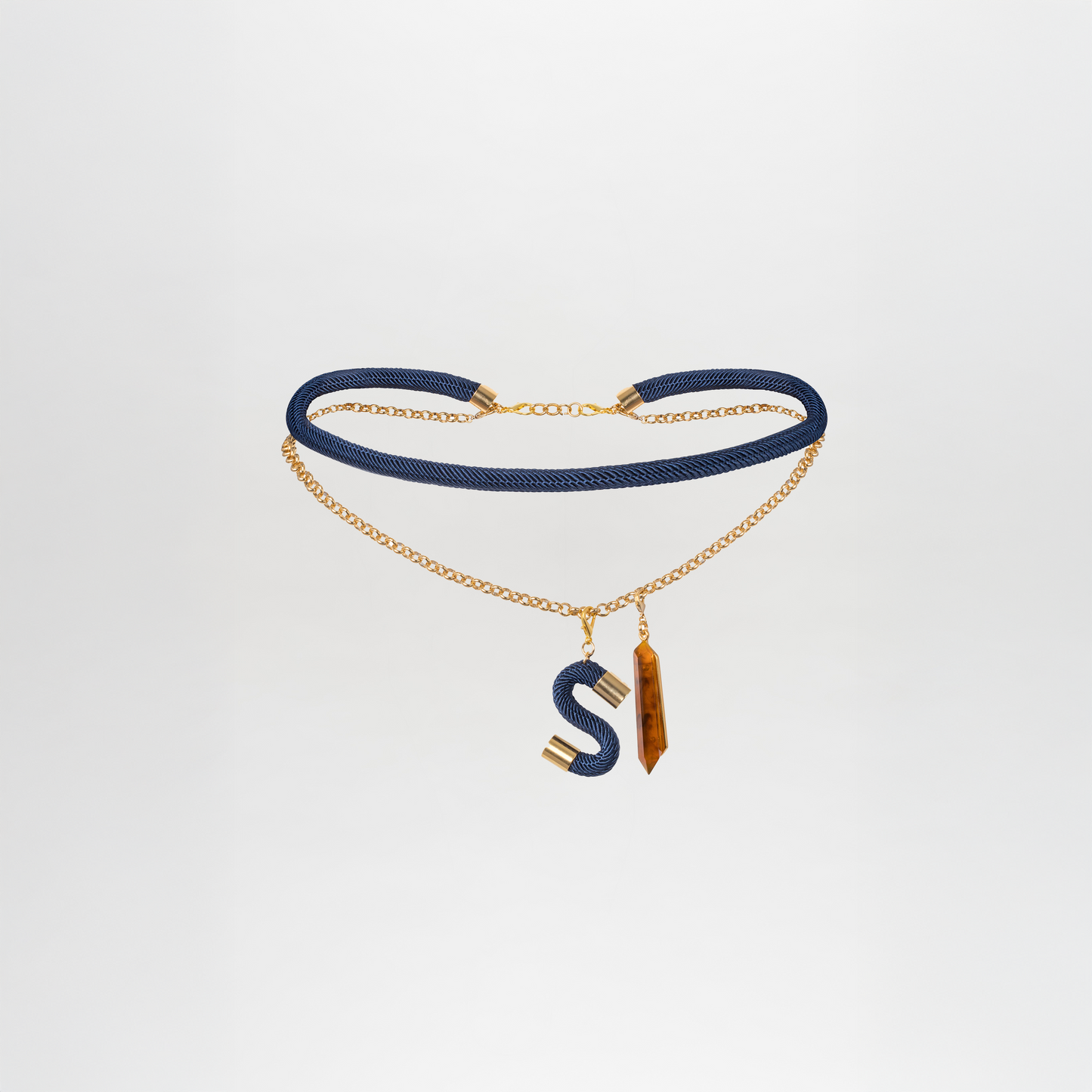 The Mireya Belt by SVT DESIGNS is a gold-plated brass chain necklace featuring a textured dark blue cord, two pendants—a large "S" and a rectangular Petralux® fantasy stone with an amber hue—shining beautifully against a plain white background.