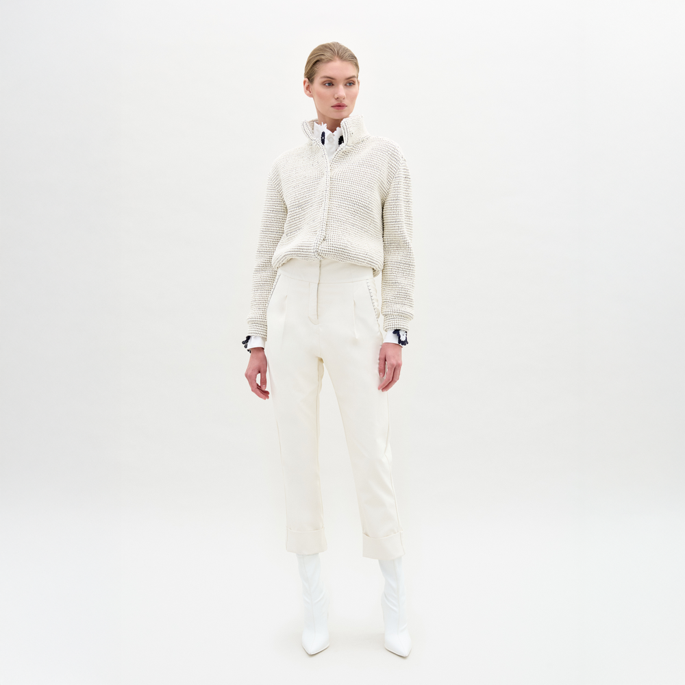 A person faces forward with hands at their sides, wearing a textured white sweater over a collared shirt, SVT DESIGNS Moad Pant—high-waisted and cropped in Italian fabric—and white pointed boots against a plain white background.