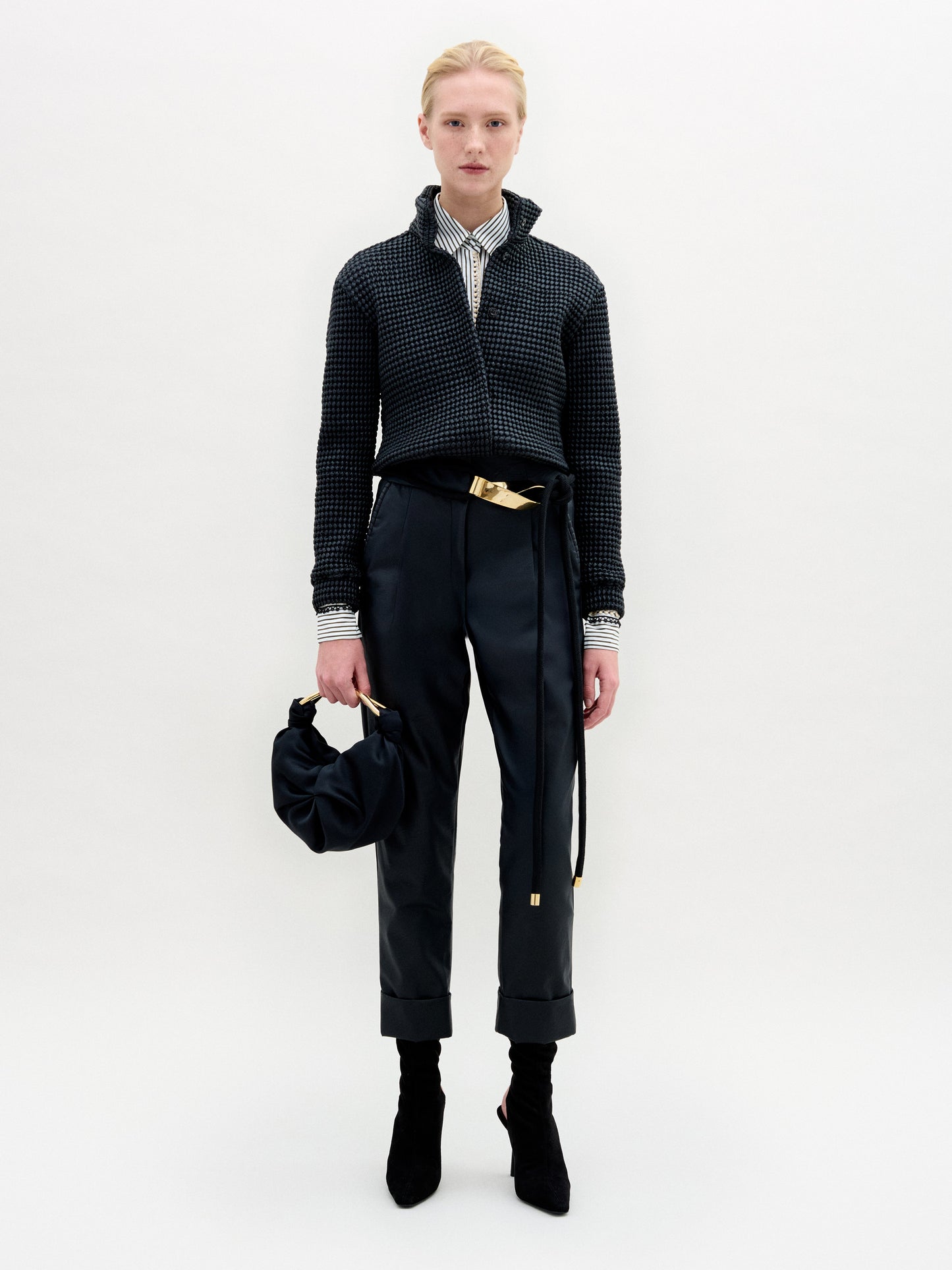 A person with blond hair stands against a white background, wearing SVT DESIGNS' Moad Pant—a high-waisted cropped style with a belt—paired with a dark jacket, striped shirt, heeled boots, and holding a dark handbag.