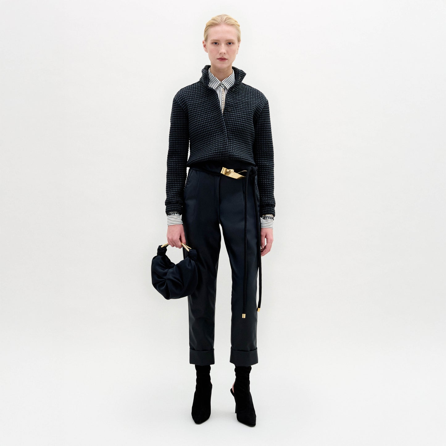A person stands against a plain white background wearing SVT DESIGNS’ Moad Pant—straight fitted black trousers crafted from fine Italian fabric—paired with a dark textured jacket, collared shirt, black boots, and holding a small black handbag.
