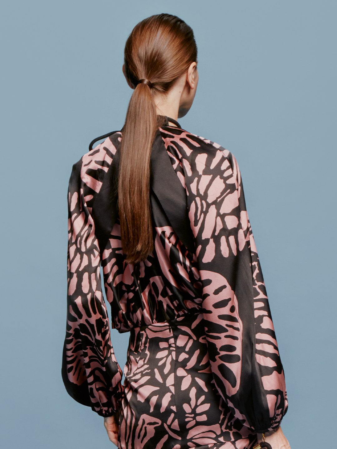 A person stands against a blue background, wearing the Moira Cape Black Pink Filigrana dress, featuring long sleeves and a high neckline, available just in time for the latest delivery schedule.