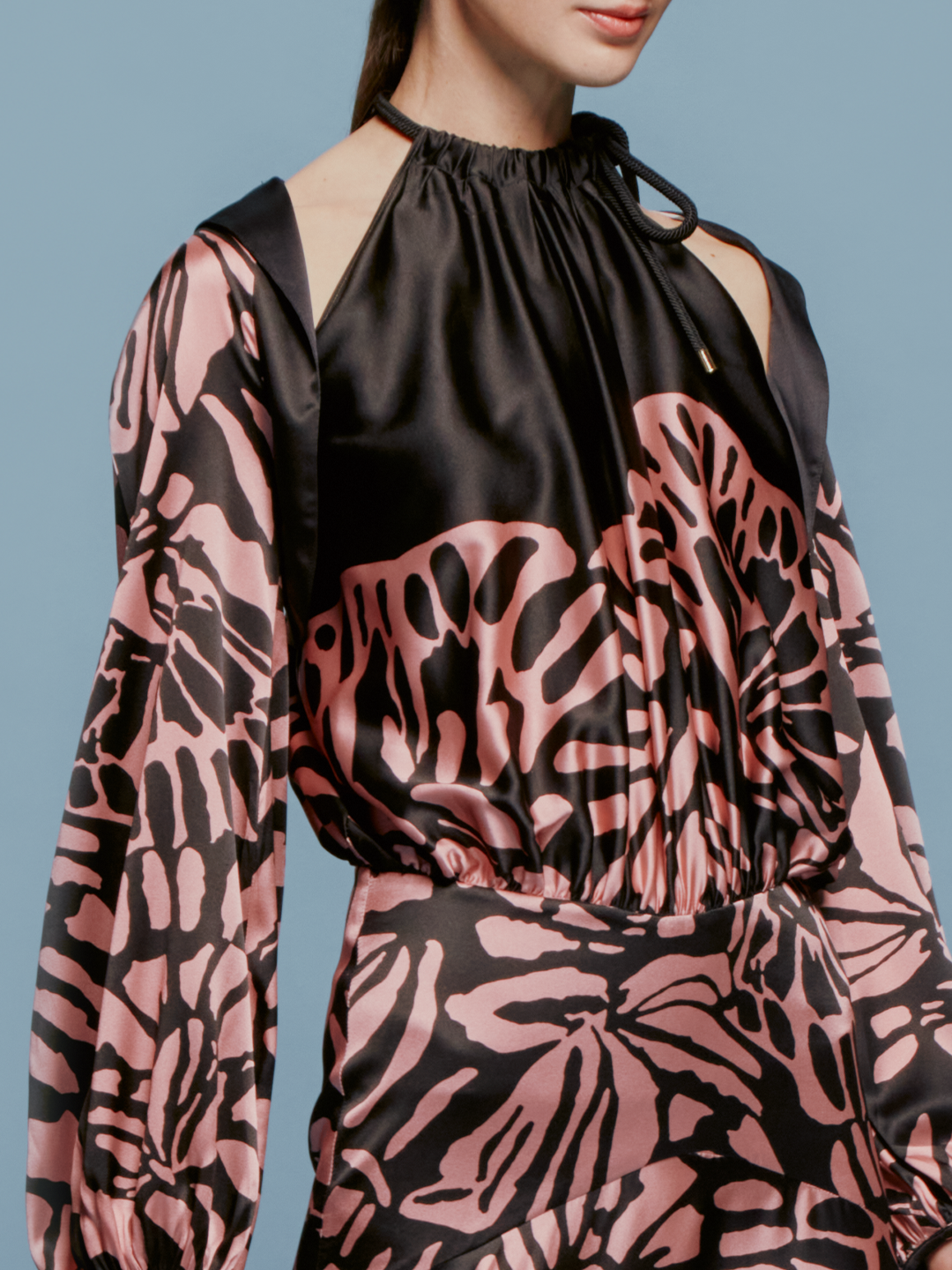A person stands against a blue background, wearing the Moira Cape Black Pink Filigrana dress, featuring long sleeves and a high neckline, available just in time for the latest delivery schedule.